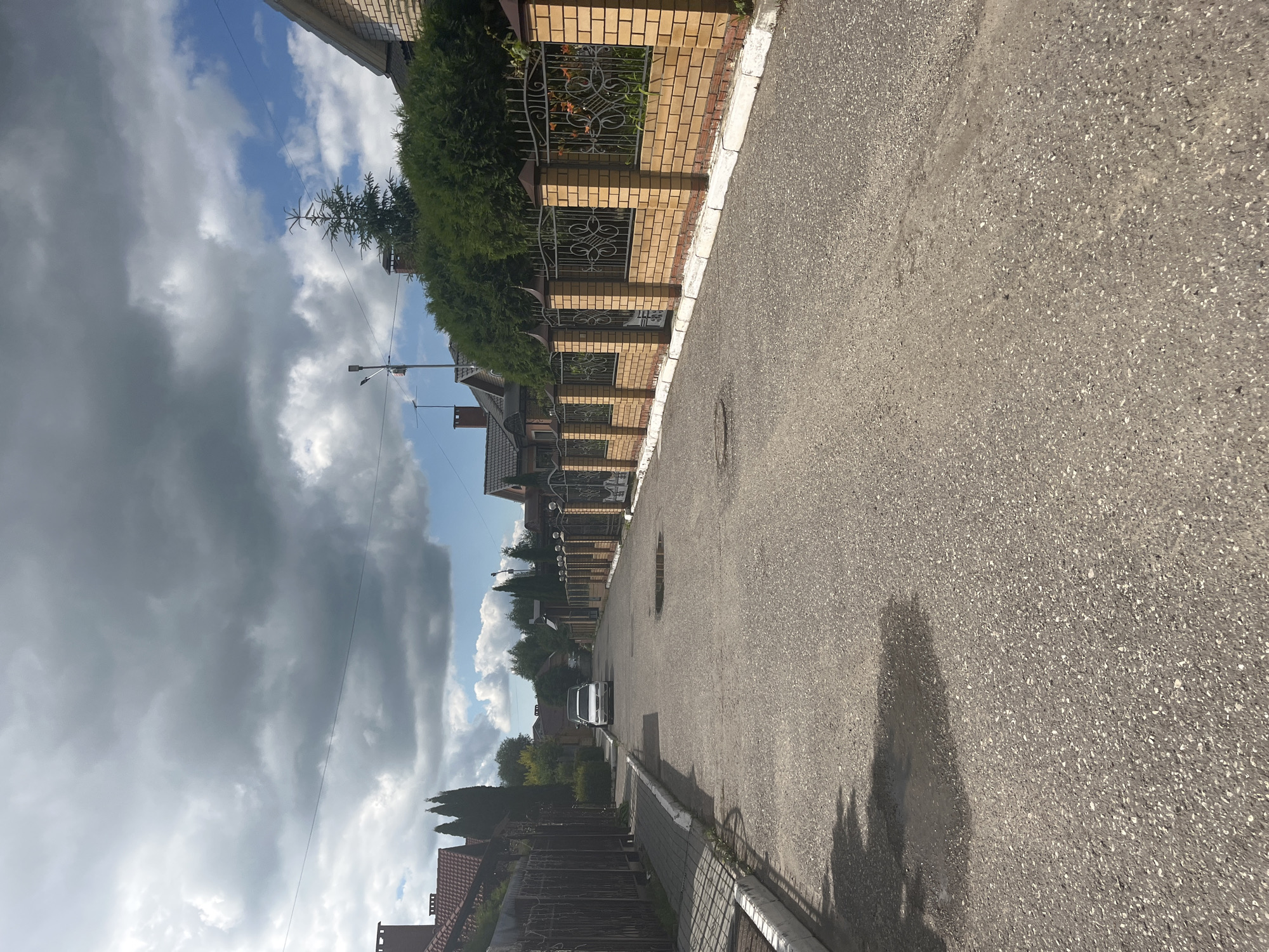 The image shows a street in the city of Orekhovka, Russia, with houses on both sides. The street is paved with asphalt and has a sidewalk on the left side. There is a white car parked on the left side of the street, and a dark SUV parked in the distance. The houses are surrounded by fences made of brick and metal. There are trees and bushes along the street. The sky is partly cloudy with some blue sky visible. The overall scene depicts a residential street in a suburban area.