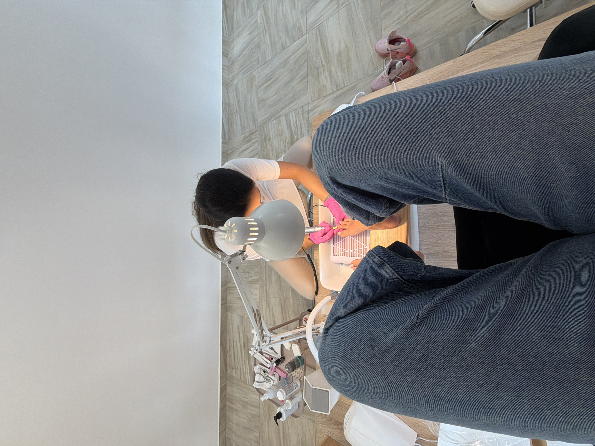 An overhead shot shows a person receiving a pedicure in what appears to be a salon. The client's legs, clad in blue jeans, are prominent in the foreground, extending towards a wooden table where the pedicure is taking place. A nail technician, wearing a white t-shirt and pink gloves, kneels at the table, working on the client's feet with a drill. Various nail care products and equipment, including a lamp and bottles, are organized on a trolley beside the technician. Pink sandals are visible on the floor to the right of the table. The flooring consists of large, light-colored rectangular tiles. The wall behind the scene is a plain, light color. There are no specific landmarks or indicators to suggest the location is in Gabrovo, Bulgaria; the scene depicts a generic nail salon environment.