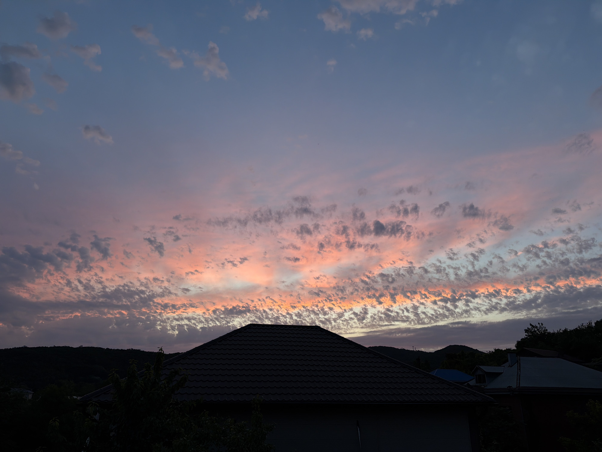 The image depicts a sunset over a residential area in Tuapse, Russia. A dark-tiled roof of a house is prominent in the foreground, silhouetted against a sky filled with scattered clouds. The clouds are illuminated with hues of pink, orange, and purple from the setting sun, creating a colorful and dramatic sky. In the background, dark hills and trees can be seen, further emphasizing the twilight setting. The overall scene is peaceful and serene, capturing the beauty of nature at dusk.