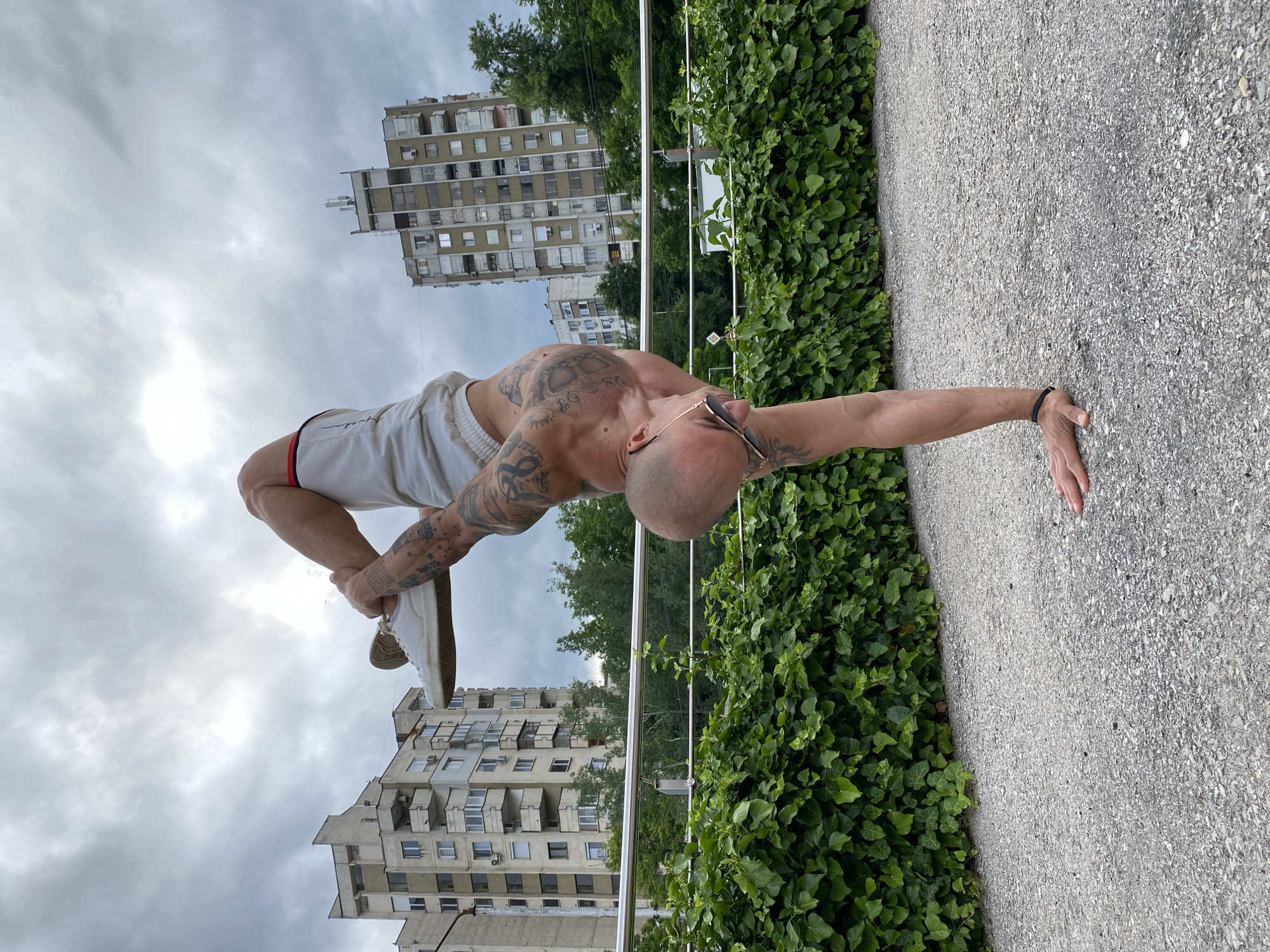 The image captures a man performing an advanced calisthenics or yoga pose outdoors in an urban setting. The man is balancing on his right arm, which is extended and firmly planted on a light grey, pebbled surface. His body is arched, with his chest and head facing upwards, and his left leg is bent, with his left hand holding his left foot. He has a shaved head, wears sunglasses, and is extensively tattooed on his torso and arms. He is shirtless and wears white athletic shorts with red and black stripes on the sides, along with white sneakers.
The setting includes a vibrant green hedge of ivy to the man's left, separating the pebbled ground from a metal railing. In the background, under an overcast sky, stand several tall, multi-story residential buildings with balconies, indicative of Eastern European architecture. Trees are visible between and behind the buildings and the hedge. The lighting suggests it is daytime, possibly under cloudy conditions. The scene reflects an urban environment consistent with locations like Pleven, Bulgaria. No legible text is visible in the image.