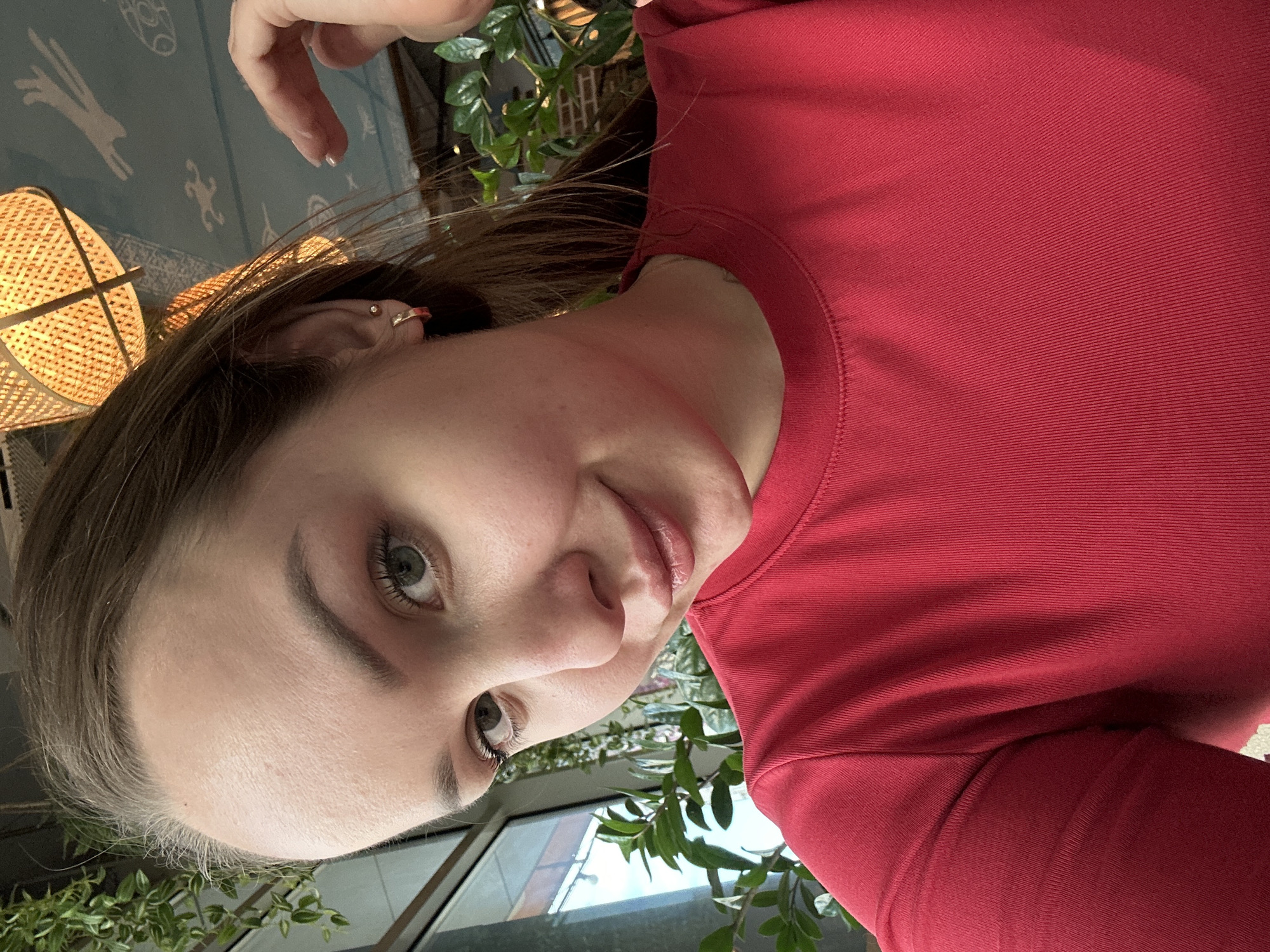 A medium close-up shot features a woman in a red top. Her fair skin contrasts with the bright color of her shirt. She has light brown hair, and her eyes are light green. A small earring is visible in her left ear. In the background, there are indoor plants and an out-of-focus patterned wall. The location in the city of Izhevsk, Russia, cannot be determined from the available information.
