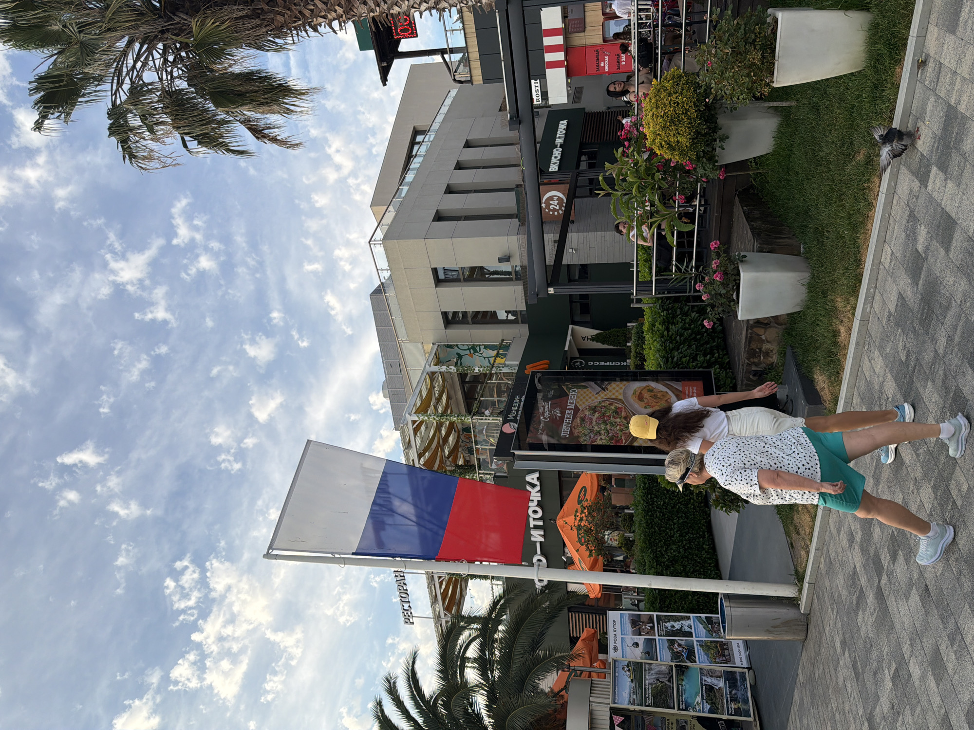 The image depicts an outdoor scene in Sochi, Russia, likely at a commercial area or promenade. In the foreground, two women are walking on a paved walkway. The woman closer to the camera is older, wearing a white and black speckled t-shirt, turquoise shorts, and light blue sneakers. She is looking to her right. The second woman, younger, is wearing a white t-shirt, a yellow baseball cap, and white shorts, and she is walking ahead of the first woman. A Russian flag is prominently displayed on a flagpole to the left of the frame.
Behind the women, there is a multi-story building with several businesses on the ground floor. Signs for "РЕСТОРАН" (Restaurant) and "ВКУСНО-И-ТОЧКА" (Vkusno i tochka, a Russian fast-food chain) are visible. Palm trees are present, suggesting a warm climate, consistent with Sochi's location on the Black Sea coast. Several outdoor seating areas with tables and chairs are visible, some with umbrellas. Planters with flowers and greenery are placed around the area, and a pigeon is seen on the ground in the lower right quadrant of the image. The sky is partly cloudy with a bright, diffused light, indicating it is daytime. The overall atmosphere appears to be casual and relaxed, consistent with a public recreational or dining space.