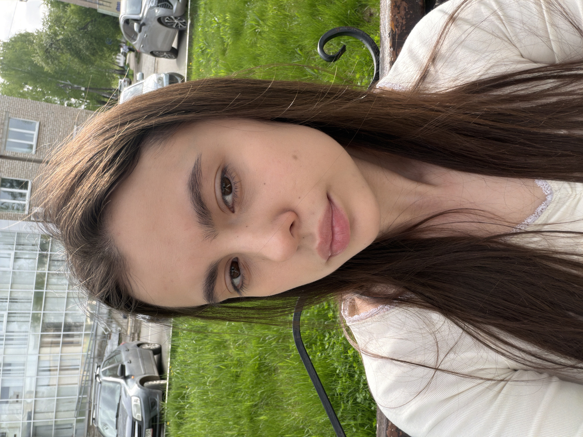 A young woman with long, dark brown hair is sitting on a bench outdoors. She is wearing a white long-sleeved shirt with lace trim at the neckline. The woman is looking at the camera with a slight smile.
Behind her is a grassy area and a building with a grid of windows. Cars are parked in the background. The location is potentially in Chaikovsky, Russia, given the prompt context. A curved metal piece, part of the bench, is visible.