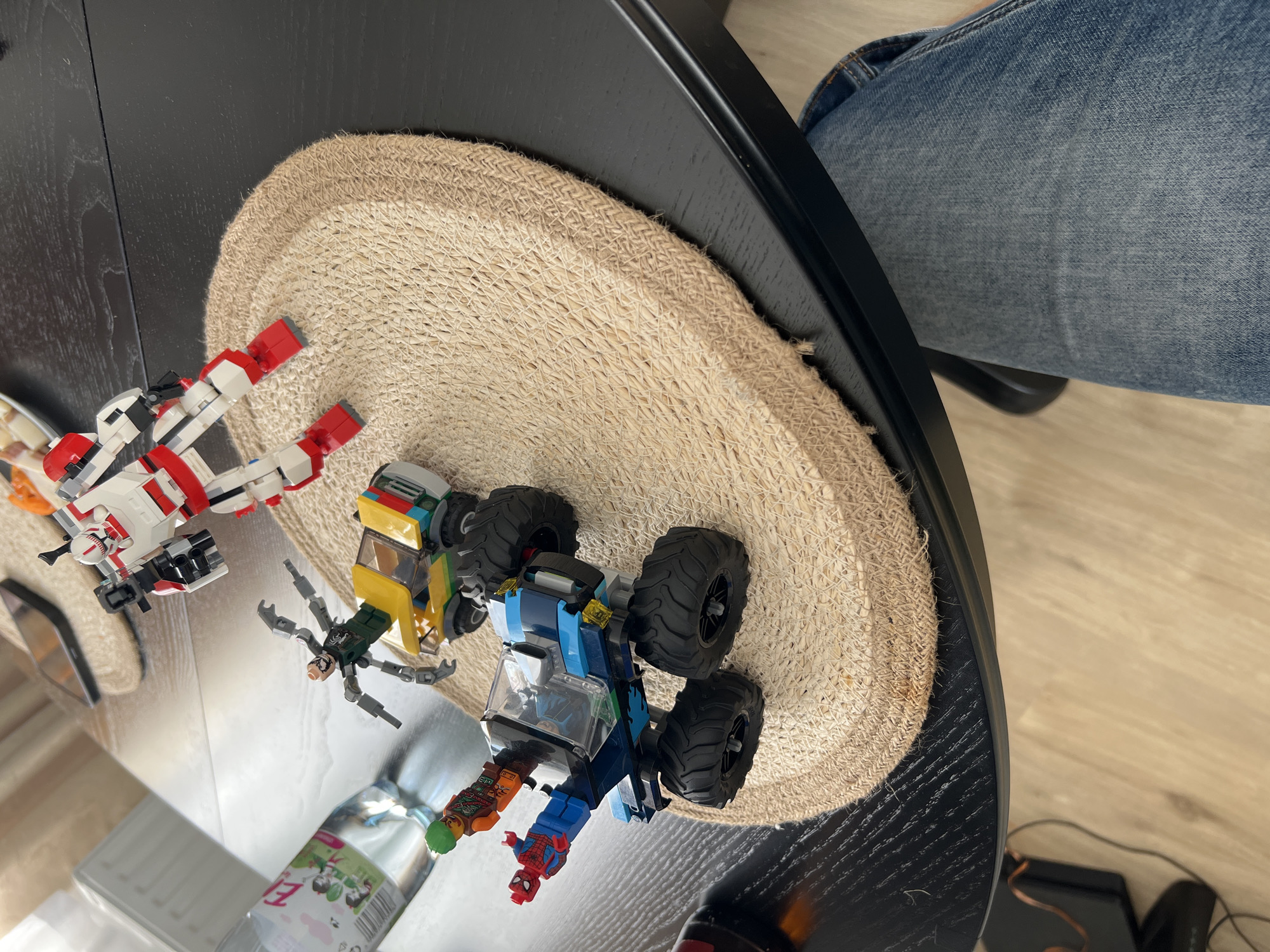A Lego action scene is staged on a woven placemat on a dark wood table. In the foreground, a blue and green Lego monster truck with four large wheels carries two minifigures: one dressed as Spider-Man and another in an orange and green outfit. A dark grey, multi-limbed Lego creature is perched on top of the truck. Behind the truck, a yellow and red Lego vehicle is parked. In the background, a white and red Lego robot stands upright. A clear plastic bottle of water, with a pink and green label, is positioned to the left of the scene. The image is taken from a high-angle perspective, looking down at the table. The background includes a blurry view of a white radiator and a smartphone on a surface. The overall setting suggests an indoor environment. No specific location in Budapest, Hungary, can be identified from the visual information provided.
