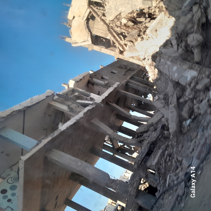 The image presents a devastated urban scene in Khan Yunis, Palestinian Territory, under a clear blue sky. Multiple multi-story concrete buildings exhibit severe structural damage. On the left, a large concrete structure stands partially collapsed, revealing numerous fractured and bent support pillars and exposed concrete slabs across multiple levels. Extensive debris, including fragmented concrete, dust, and twisted rebar, covers the ground level around and within this building. To the right, another building appears more completely destroyed, reduced primarily to a large mound of rubble and broken concrete with exposed rebar. No individuals, ongoing actions, or interactions are visible within the scene, which is characterized by static ruin under bright daylight conditions.