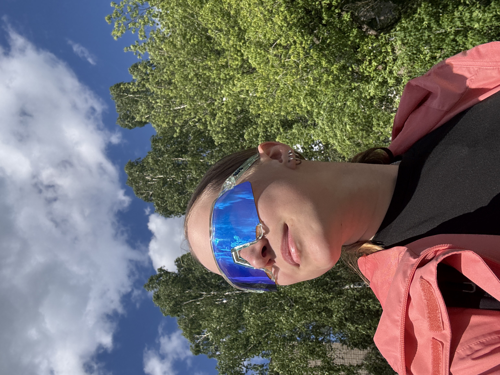 A portrait of a person wearing futuristic blue-tinted sunglasses is set against a backdrop of a tree and sky. The person, dressed in a black shirt and coral-colored jacket, is facing slightly away from the camera. The sky is partly cloudy, with blue patches visible. The tree's green foliage fills the background. The location is not discernible.