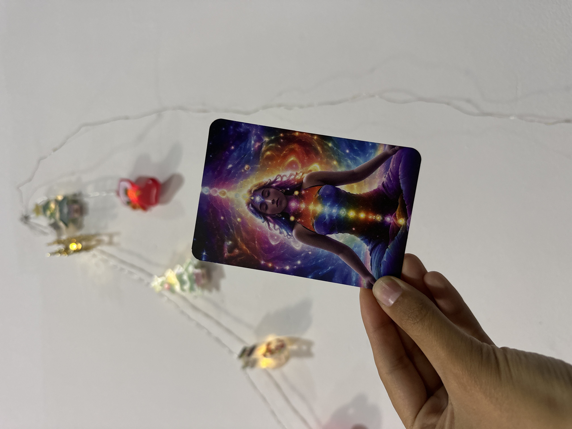A hand holds a card depicting a woman against a cosmic background. The woman has her eyes closed, and light appears to be emanating from her forehead and chest. The background of the card features a colorful nebula. The card is being held over a white surface with a string of lights and decorative elements. The location is not identifiable as Da Lat, Vietnam from the provided information.