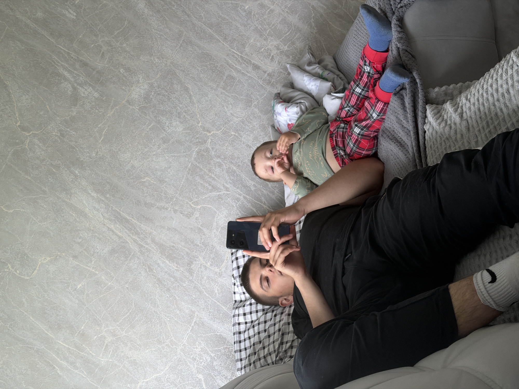 Two males are lying on a couch. On the left, a young male with short hair is wearing a black t-shirt and black pants. He is holding a black smartphone in his hands and looking at it. On the right, a toddler is wearing a light green long-sleeved shirt with a dinosaur pattern and red plaid pants. He is lying on his back with his hands near his face. The couch is covered with blankets and pillows. The background is a grey textured wall. The image appears to be taken indoors, possibly in a residential setting. There is no identifiable information to place the image in Andreyevo, Russia.