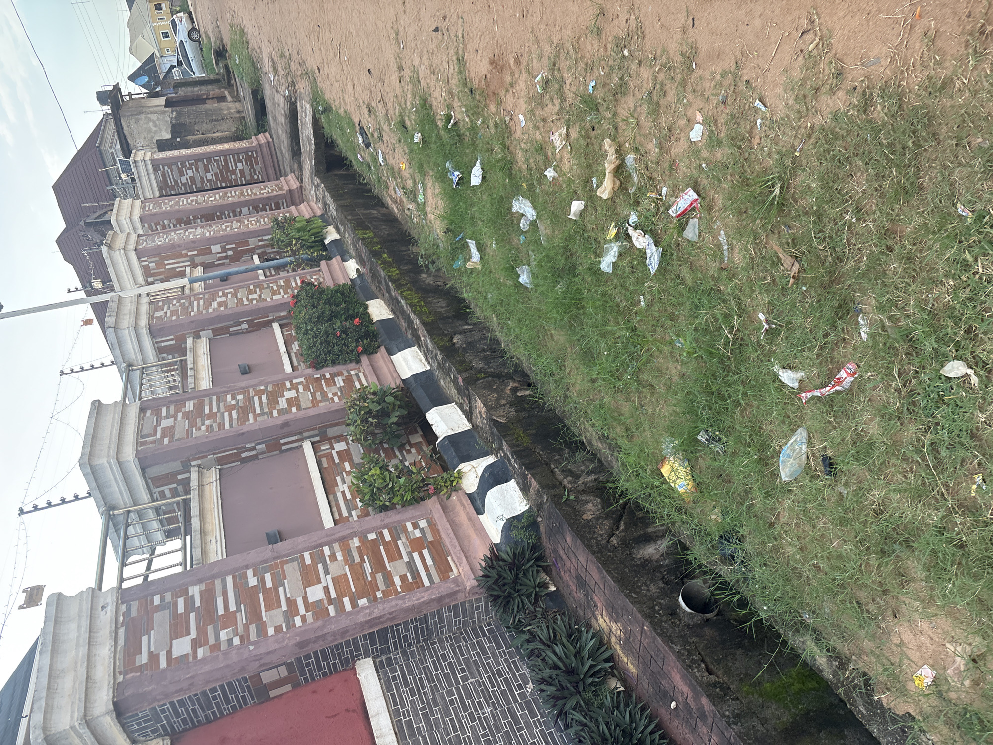 A street in Obosi, Nigeria, is depicted with a sidewalk adjacent to a street and a concrete drainage ditch. The ditch is filled with debris and litter, including plastic bags, wrappers, and bottles. The sidewalk is also littered with trash. Buildings are visible on the other side of the sidewalk, including a house with a decorative brick wall and fence. The sky is overcast.