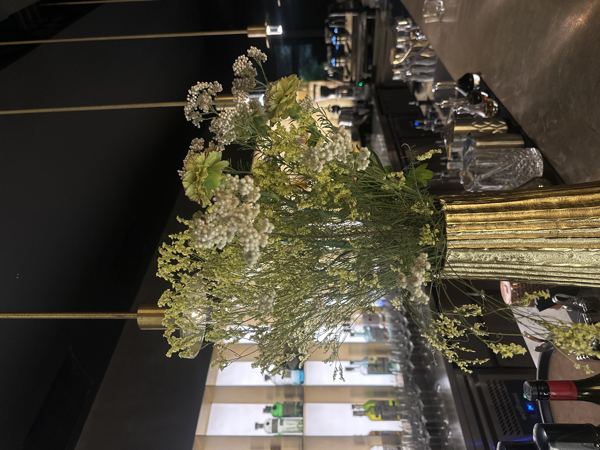 The image shows a floral arrangement in a golden vase on a bar top. The flowers are predominantly white and light green. Behind the arrangement, there is a blurred view of bar equipment, glassware, and liquor bottles on illuminated shelves. The location is likely in Sofia, Bulgaria, but there is no specific evidence in the image to confirm this.