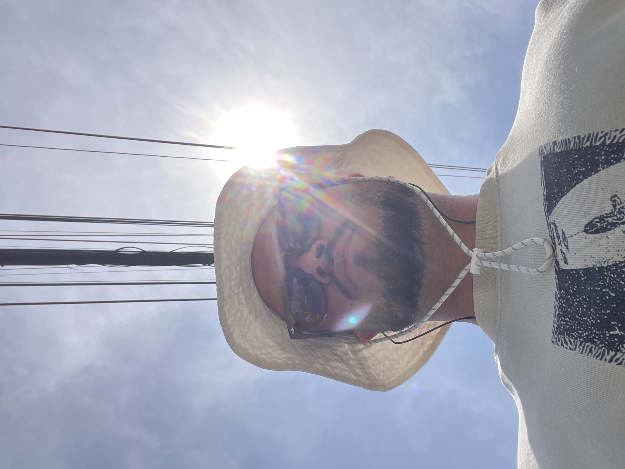 An image, rotated ninety degrees counter-clockwise, depicts a person from an upward-looking perspective against a bright, hazy sky. The sun is prominently visible in the upper left quadrant, causing significant lens flare and backlighting the subject.
The person wears a light-colored, possibly straw or fabric, wide-brimmed hat, and dark sunglasses. A beard is visible beneath the sunglasses. They are dressed in a light-colored t-shirt with a large dark graphic print on the right side; the partial word "AMAZING" is discernible within the print, along with other abstract patterns. A white and black cord, possibly a drawstring for the hat or a headphone cord, is visible around their neck.
Several horizontal utility lines cross the upper portion of the frame, originating from the left. The sky is a light blue with subtle cloud formations, indicating clear weather. The strong sun suggests it is midday or early afternoon. No specific location landmarks are visible, placing the scene in an unknown outdoor setting. The camera appears to have been held low, looking up at the subject.