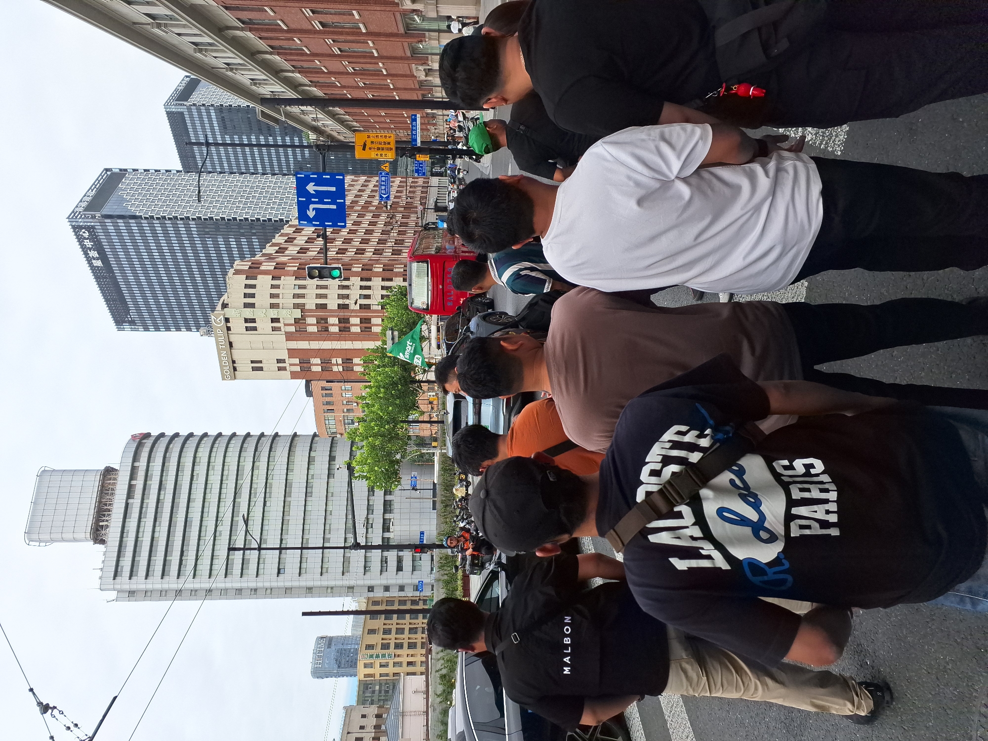 The image shows a street scene in Hongkou District, China, with several people standing on a crosswalk. In the foreground, a group of men are viewed from behind, some wearing t-shirts with logos. A black t-shirt with "MALBON" in white lettering is visible. Another black t-shirt displays "LACOSTE R.Loco PARIS" in white and blue lettering. Behind the pedestrians, cars and motorcycles are on the street, and a red bus is visible. Tall buildings line the street, including a modern silver skyscraper with a rectangular top section and a darker skyscraper with a patterned facade. Traffic lights and street signs are present. The sky is overcast.