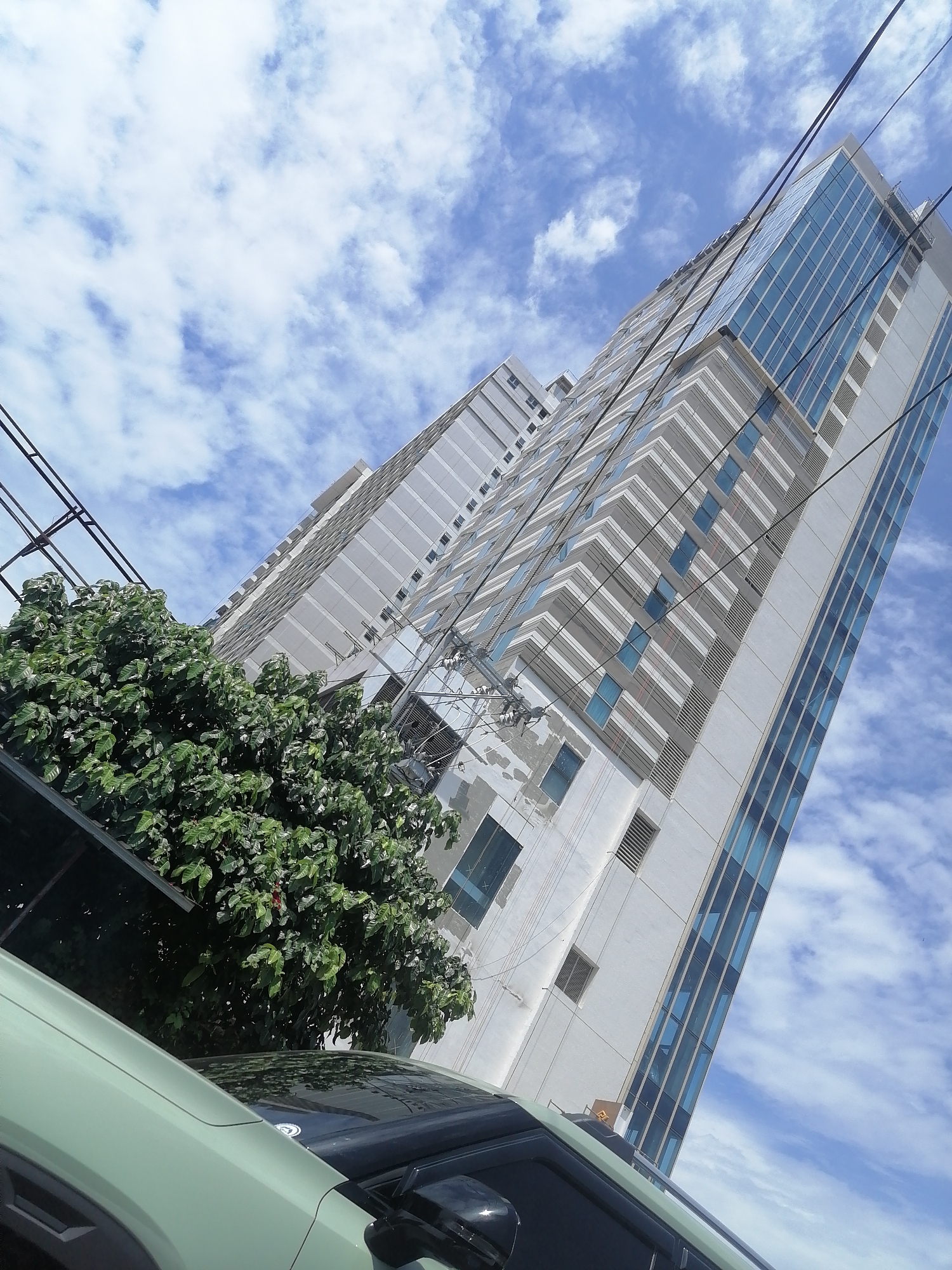 This media file presents a low-angle, upward-looking view of a modern high-rise building against a clear blue sky dotted with scattered white clouds, indicative of a bright, sunny day in Davao City, Philippines. The prominent building, likely a residential or office complex, features a contemporary design with large sections of reflective blue-tinted glass windows and gray and white striped facades. A less detailed, similar building is partially visible in the background to the left.
In the foreground, a dense, vibrant green tree occupies the lower left portion of the frame, partially obscuring the building. The front-left section of a light green vehicle, possibly an SUV, is visible at the bottom of the image, suggesting the photograph was taken from street level. Several black utility lines or power cables crisscross diagonally across the sky and the building, adding an urban characteristic to the scene. There are no people visible, nor is any discernible text present on the buildings or vehicle. The image captures a static moment of urban architecture under fair weather conditions.