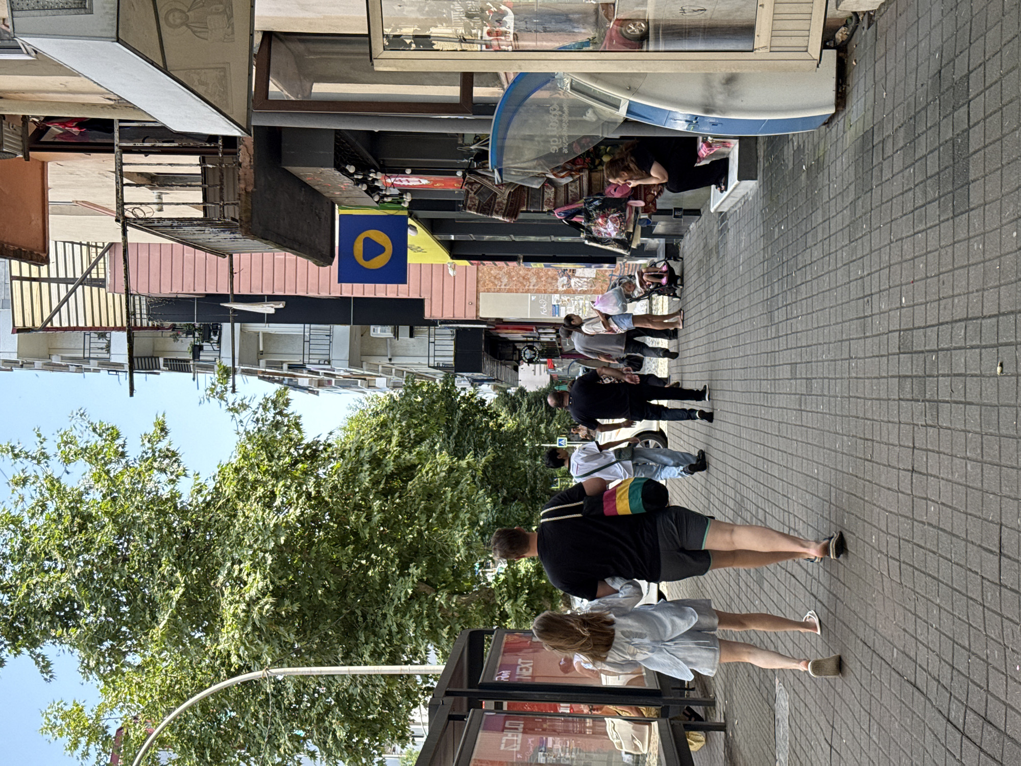 The image shows a street scene in an urban environment. A paved sidewalk is lined with buildings on the right and a bus stop on the left. Several people are walking on the sidewalk. In the foreground, a couple walks away from the camera, holding hands. The man wears a black t-shirt and shorts, and the woman wears a light blue dress and sandals. Behind them, other pedestrians are visible, including a person in a white t-shirt and jeans, and a person in a black t-shirt and black pants. Shops are located along the right side of the street, displaying merchandise such as rugs and bags. A woman sits on a step outside one of the shops, looking to the side. A baby stroller is parked near the shops. Trees with green foliage line the left side of the street, partially obscuring the view of the buildings behind them. The sky is clear and blue.