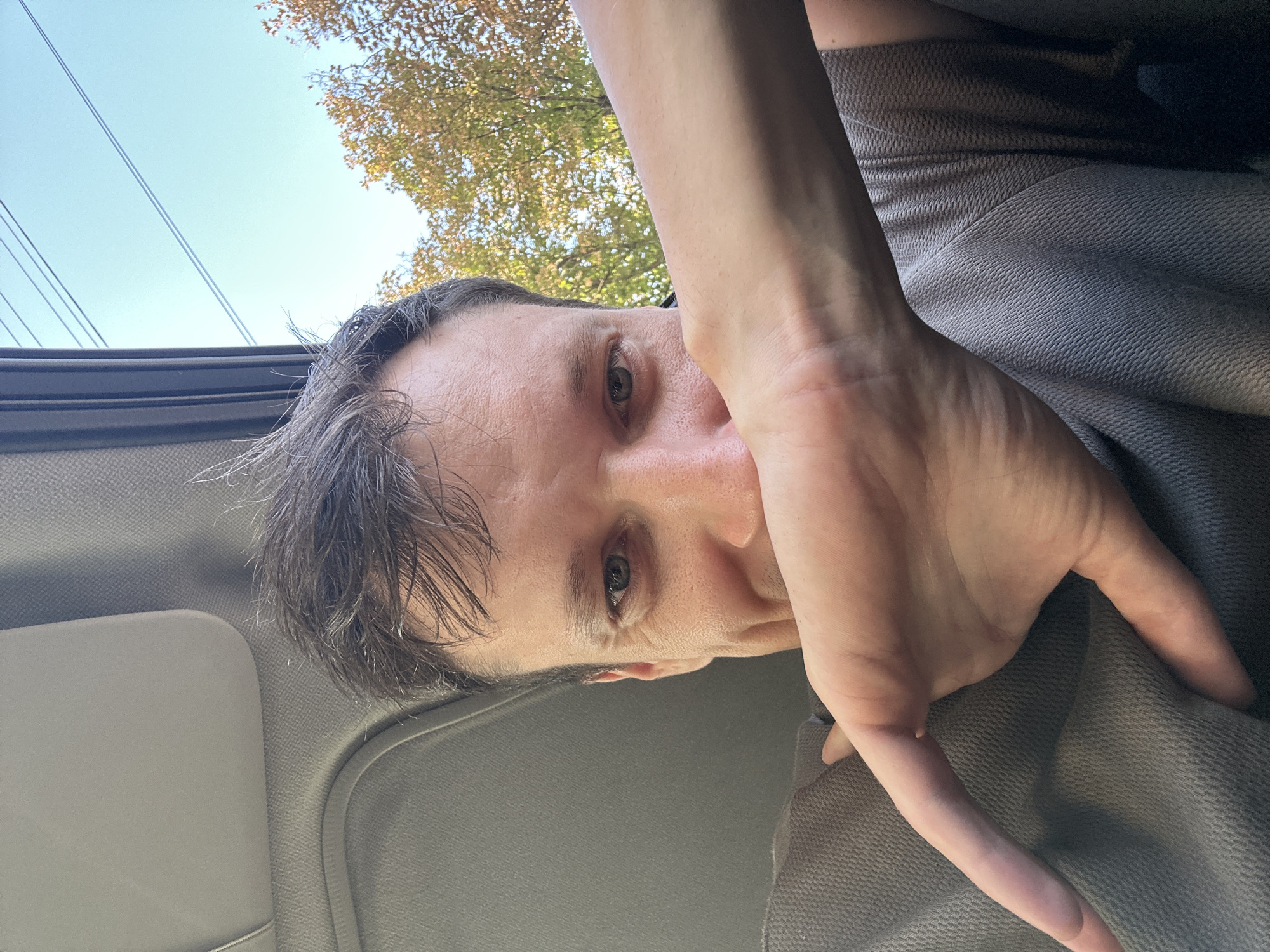 This is a selfie taken from inside a vehicle, possibly a car or bus, with the user's face partially obscured by their hand and arm. The background shows a clear blue sky with some power lines and autumn foliage from trees. The interior of the vehicle is visible with a gray fabric-covered surface, possibly a headliner or sun visor. The man appears to be looking towards the camera, with his left eye visible. The image does not provide any specific details that would indicate the location in the city of Kamianets-Podilskyi, Ukraine.