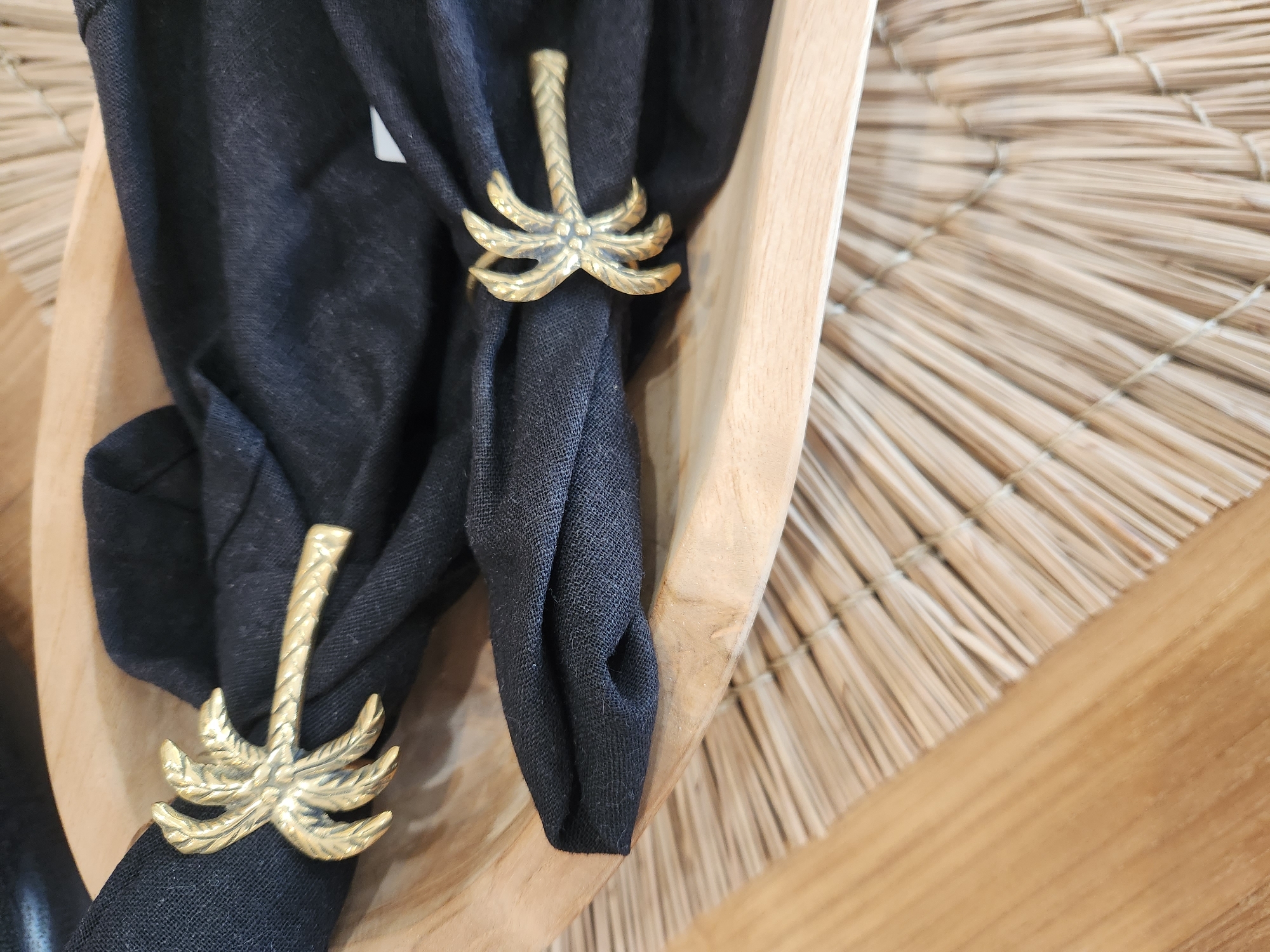 The image shows a close-up of two black linen napkins folded and secured with gold palm tree-shaped napkin rings. The napkins are resting in a light-colored, shallow wooden bowl, which is placed on a woven straw placemat. The placemat is on a wooden surface. The scene appears to be an indoor setting, possibly a dining table. The city of Maitland, United States of America is not depicted in the image.