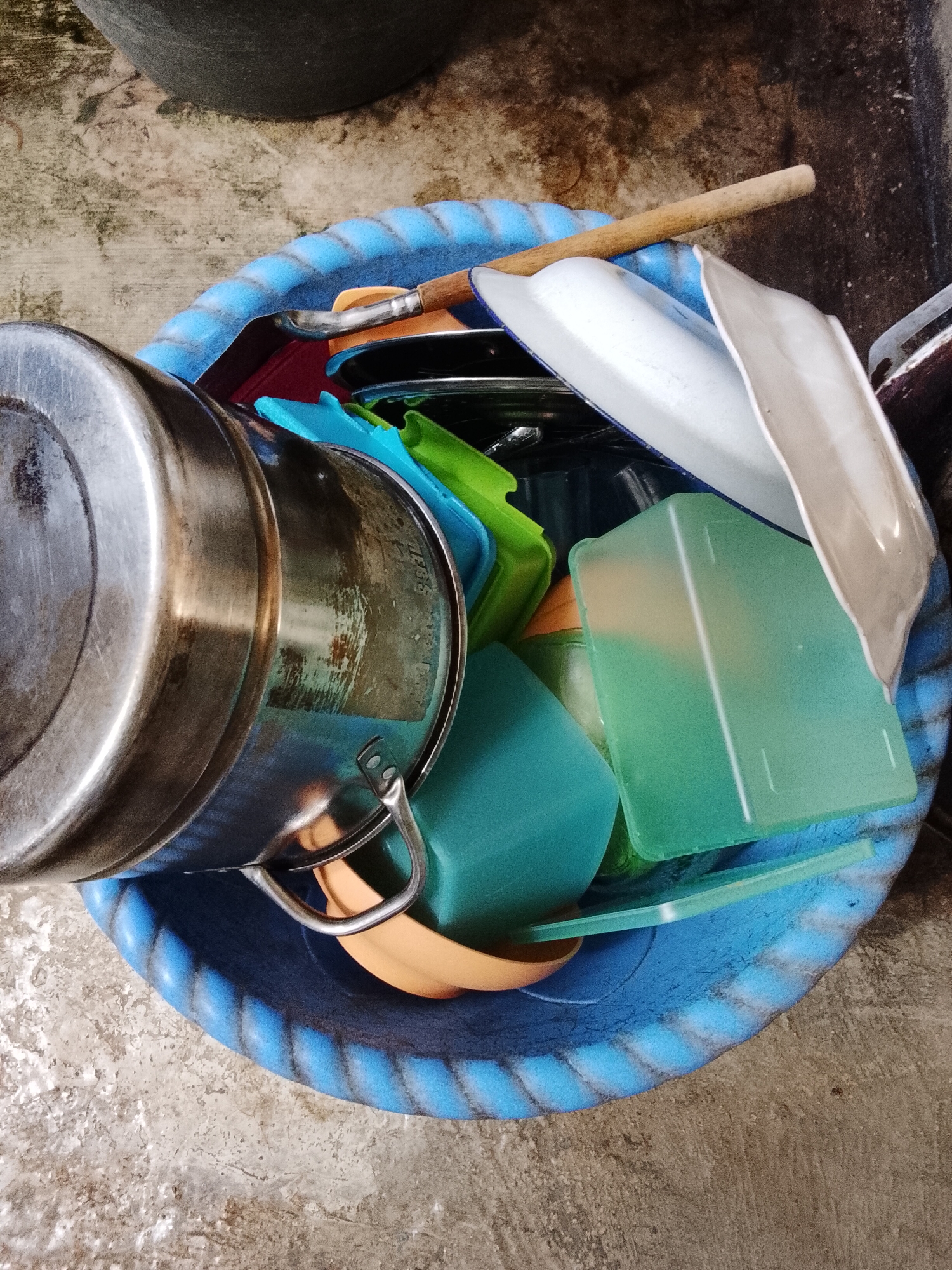 A blue basin, filled with various kitchen items and cleaning tools, sits on a concrete surface. The basin is textured with a rope-like pattern around its rim. Inside, a large metal pot with a handle is prominently displayed, lying on its side. Other objects include several green and orange plastic containers, white plates, and a wooden-handled brush. The surrounding environment appears to be an outdoor space, possibly a yard or alley, with a rough concrete ground and a darker, possibly metallic, structure in the background. The scene suggests a domestic setting where cleaning or storage of kitchenware is taking place.