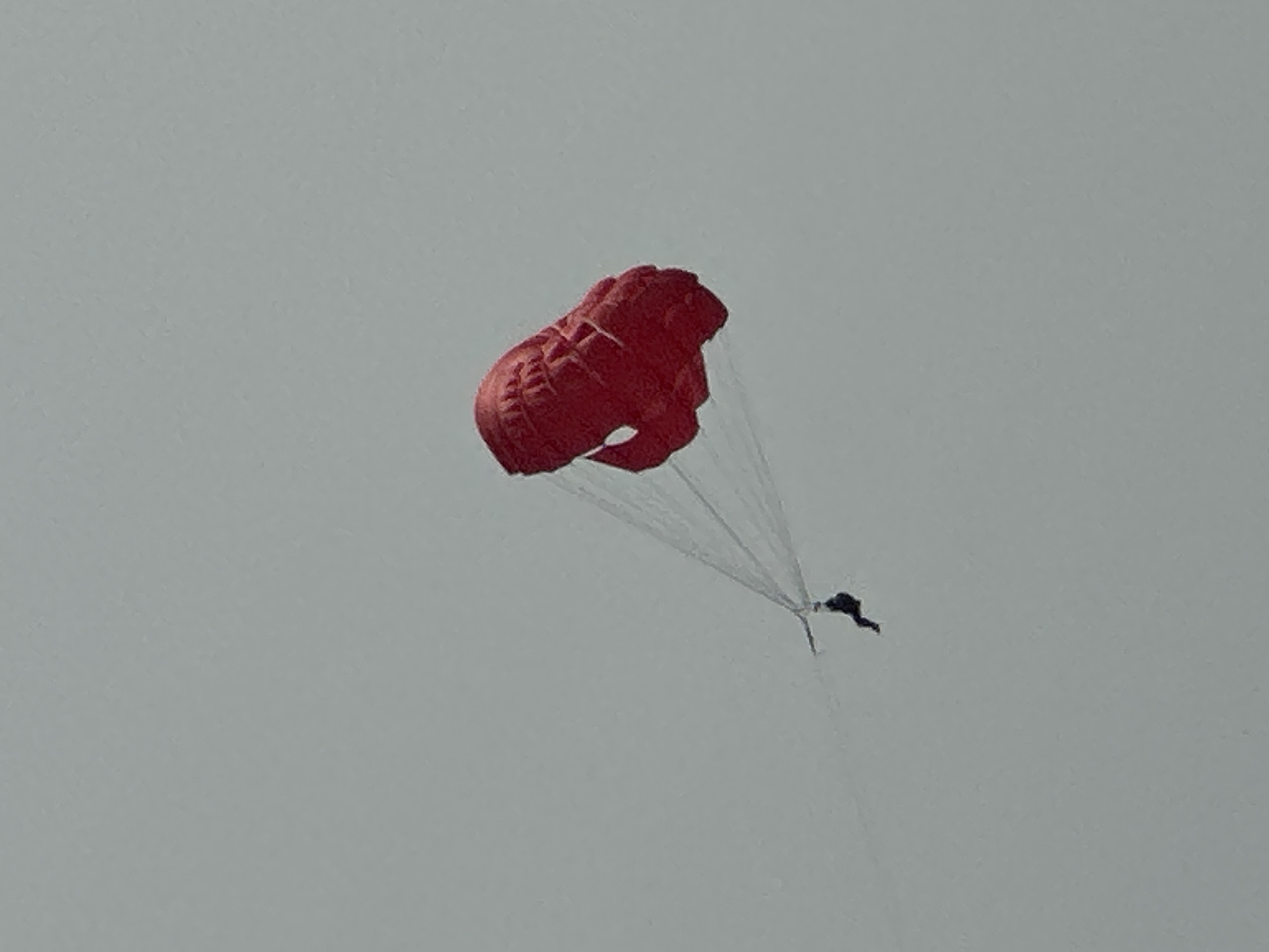 A paraglider, wearing a dark suit, descends in a red parachute against a pale gray, overcast sky. The parachute is deployed and appears to be functioning normally, with a few pleats visible in its fabric. The paraglider is suspended below the parachute by white cords, and their body is positioned in a relaxed posture. The scene is an aerial view, with no discernible landmarks or context to indicate a specific location within Boreti, Montenegro. The overall atmosphere is one of peaceful descent in an expansive, featureless sky.