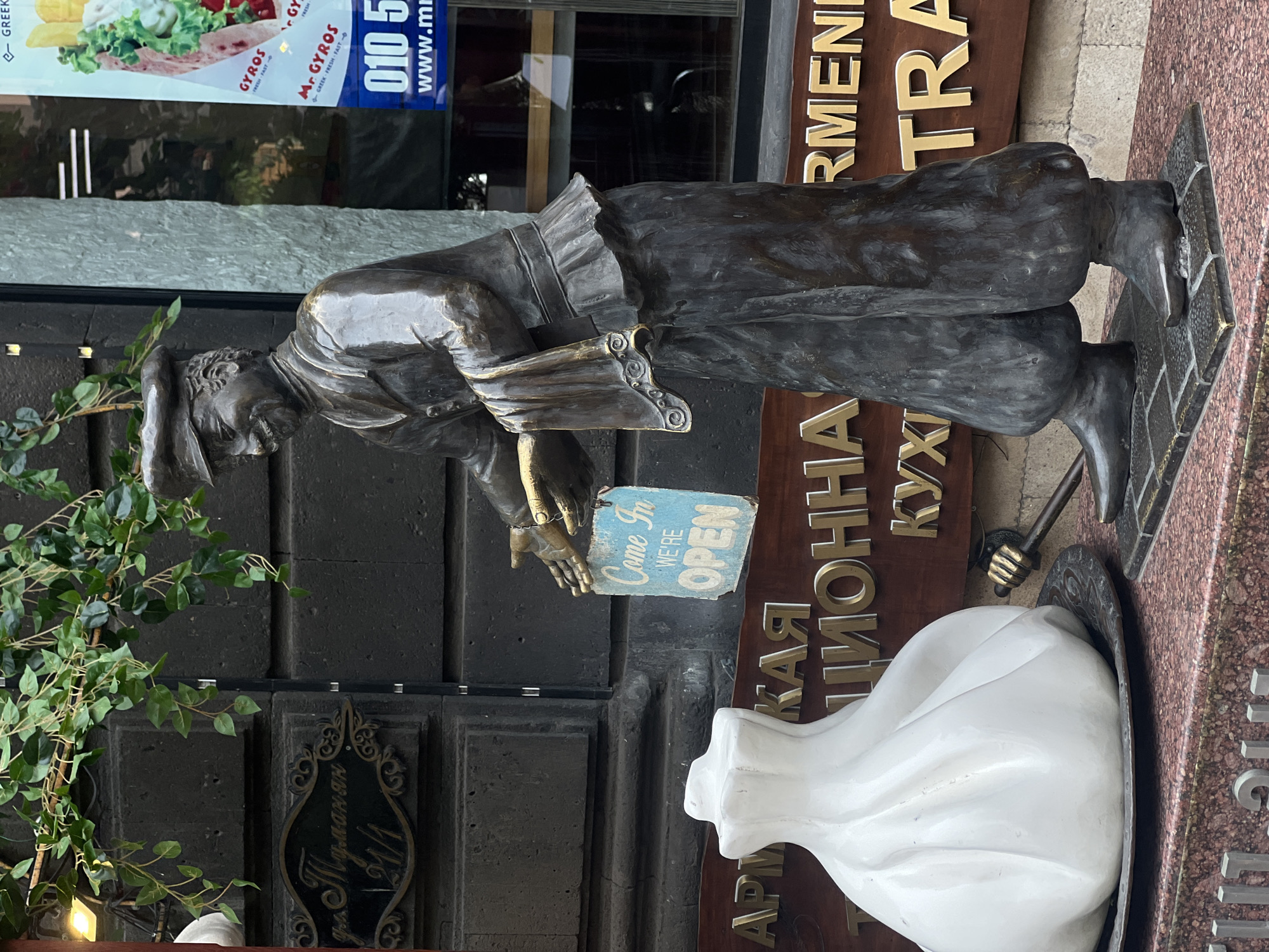 The image shows a bronze statue of a man holding an "Open" sign in front of a restaurant. The restaurant has signage in multiple languages including Armenian, and Russian. The statue is located on a street in Yerevan, Armenia. The restaurant also appears to offer Greek gyros.