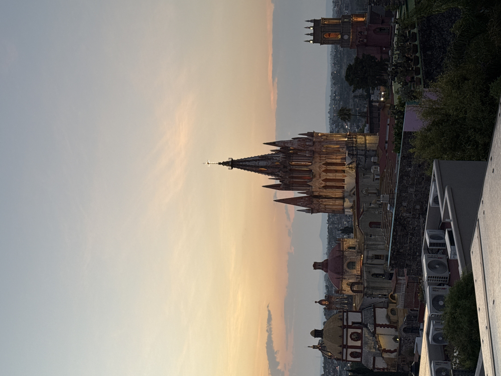 The image captures a cityscape in San Miguel de Allende, Mexico, during what appears to be twilight. The skyline is dominated by the iconic towers and spires of the Parroquia de San Miguel Arcángel, the city's main church. The church is lit by the setting sun, casting a warm golden glow on its facade. Several other buildings, including domed structures, are visible below the church.
The foreground shows elements of modern construction, possibly a rooftop terrace or balcony, including air conditioning units. Greenery is present, contrasting with the urban environment. The sky is a gradient of colors, transitioning from a light blue to orange and yellow near the horizon.