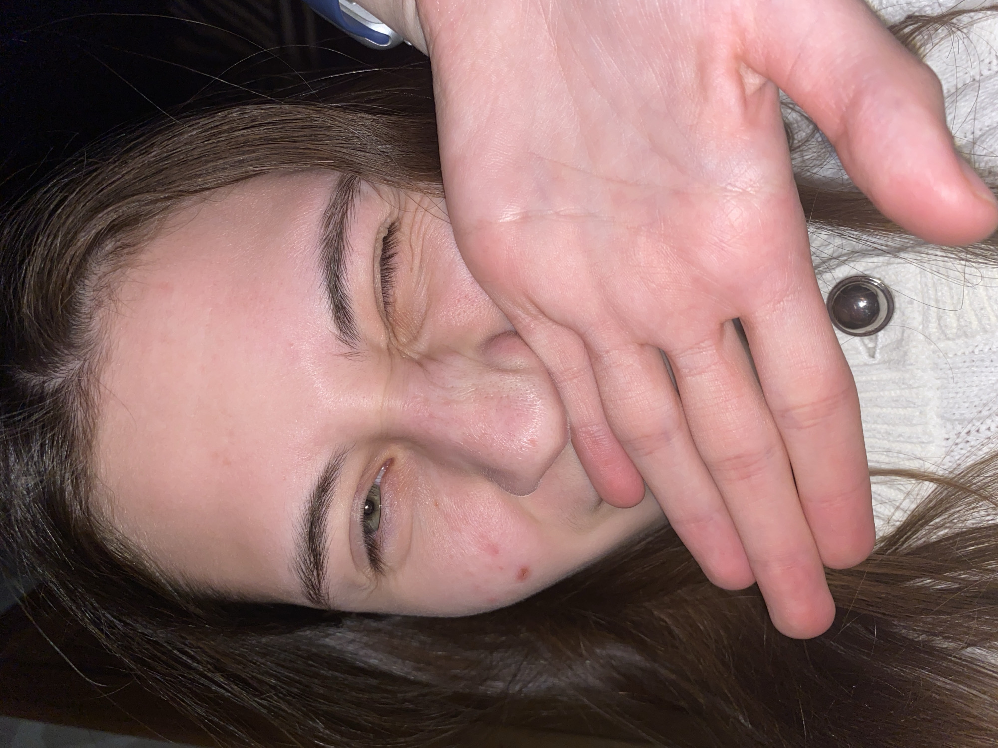 A close-up, angled portrait shot shows a person, likely a young woman, lying down indoors. Her head is tilted, and her long, brown hair spreads across the frame and partially over a light-colored, knitted garment, possibly a cream or white sweater or cardigan, which features a prominent metallic button near the right side of the image.
Her hand, with fingers slightly curled, covers her mouth and part of her nose. One of her eyes, appearing green or hazel, is visible and open, with a slight crinkling at the corner that could indicate a suppressed smile or a squint. Her eyebrows are dark and well-defined. A small section of a blue and silver wrist accessory, possibly a watch or fitness tracker, is visible at the top edge of the frame. Her fair skin shows a few minor reddish blemishes on her cheek area. The background behind her head is dark and out of focus. The scene takes place in an indoor setting in Votkinsk, Russia. There is no visible text.