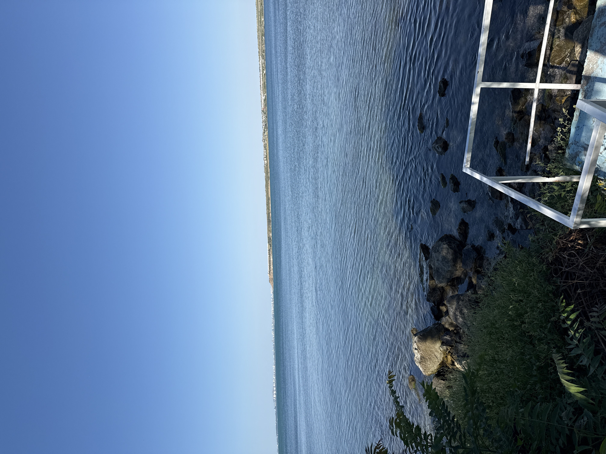The image shows a body of water with a clear blue sky above. In the distance, a city skyline is visible along the horizon. The foreground features a rocky shoreline with green vegetation and a white railing leading down some steps. The water is calm with gentle ripples, and the sunlight creates shimmering patterns on its surface. This scene appears to be a coastal area, possibly a bay or a large lake, in the vicinity of Pomorie, Bulgaria, given the characteristic landscape and presence of a settlement along the coast. There are no people or animals visible in the image.