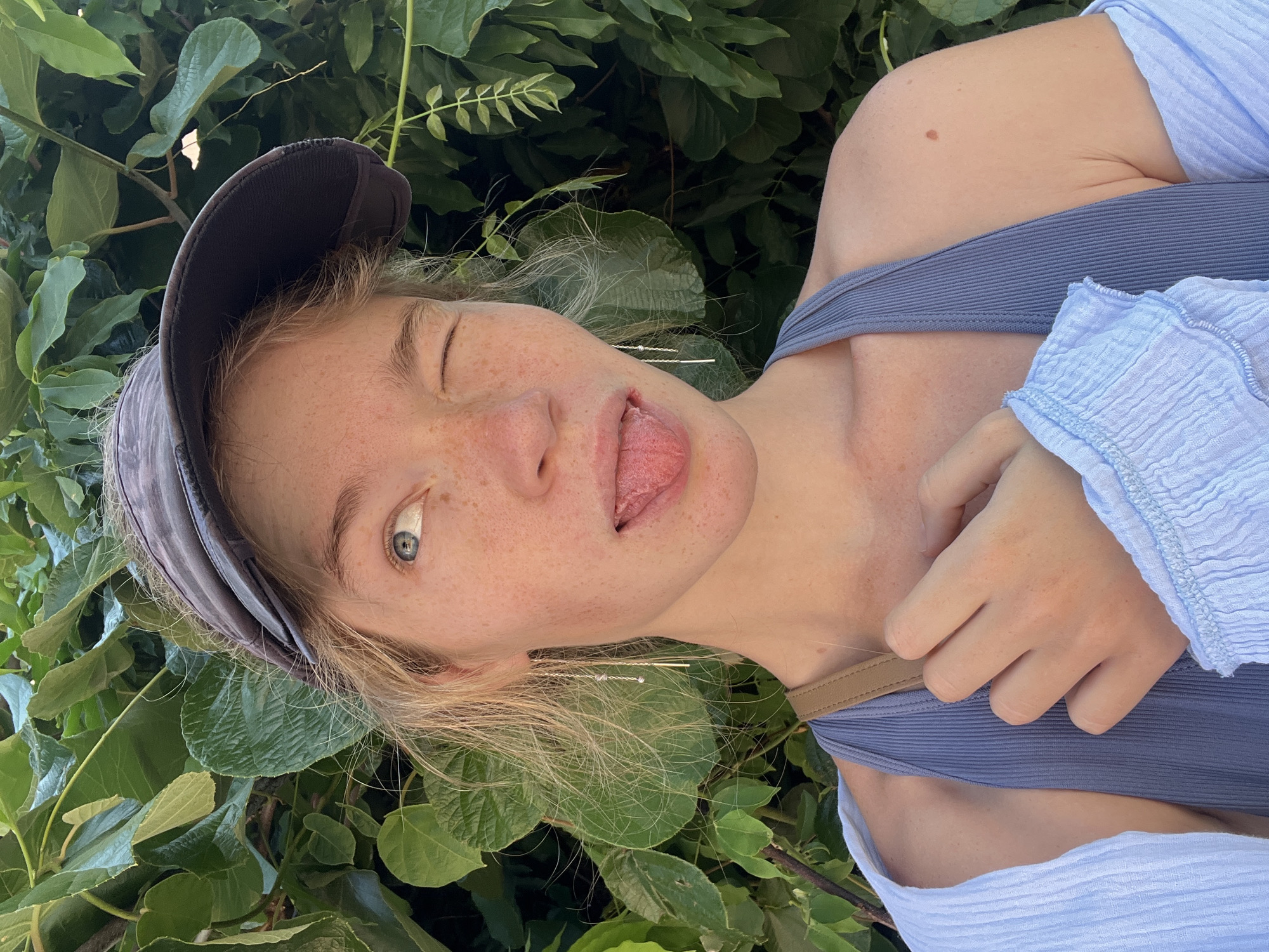 A young woman with blonde hair and freckles, wearing a dark baseball cap and a blue tank top, winks and sticks out her tongue. She is positioned in front of a backdrop of lush green foliage, suggesting an outdoor setting possibly in Budva, Montenegro. The lighting indicates a sunny day. Her left hand is visible, resting on her chest. She is also wearing dangling earrings.