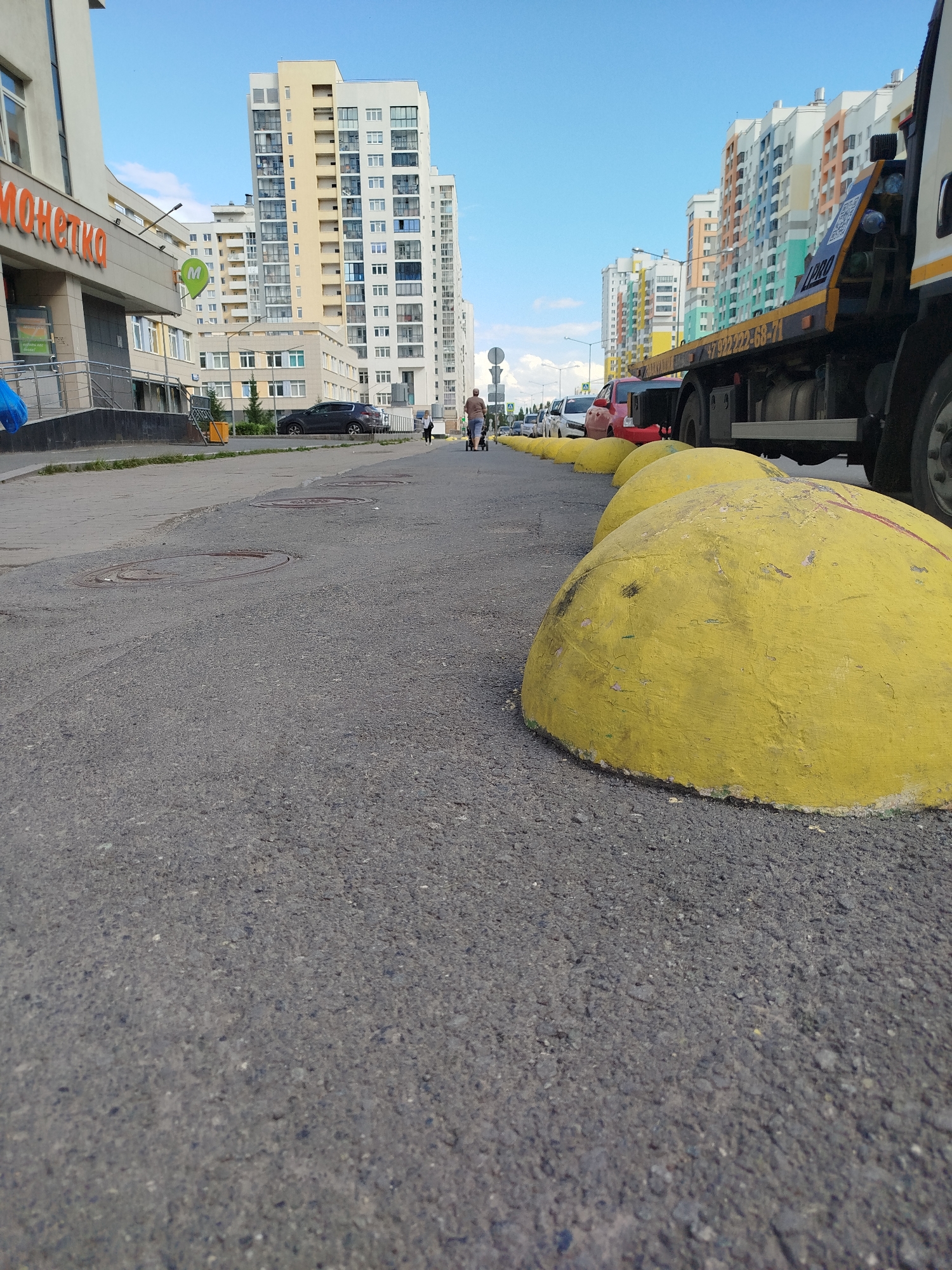 This media file depicts a street scene in Yekaterinburg, Russia, on a clear, sunny day, likely in the late afternoon or early evening given the light. The foreground is dominated by a paved asphalt surface with a row of large, bright yellow concrete bollards lining the right side of the frame. These bollards appear weathered and have some scuff marks and faded red paint on them.
The mid-ground shows a sidewalk or pedestrian area and the street. Several cars are parked along the street, including a black SUV and a red sedan. A tow truck is also prominently visible on the right side of the frame, its flatbed angled towards the viewer.
In the background, a row of tall, modern apartment buildings with colorful facades stands against a bright blue sky with scattered white clouds. The architecture suggests a contemporary urban environment.
On the left side of the frame, a building with a prominent sign reading "МОНЕTKA" in orange lettering is visible. This is likely a commercial establishment, possibly a supermarket or convenience store. A disabled access ramp leads up to the building's entrance.
Further down the street, a few people can be seen. One individual is walking with a stroller, while another person is walking in the same direction. Their presence suggests normal daily activity.
The overall impression is of a functional, everyday urban setting. There are no strong emotions conveyed, and the scene appears calm and ordinary. The visible text "МОНЕTKA" and a phone number on the tow truck "+7 (922) 222-68-71" provide specific location and context clues.