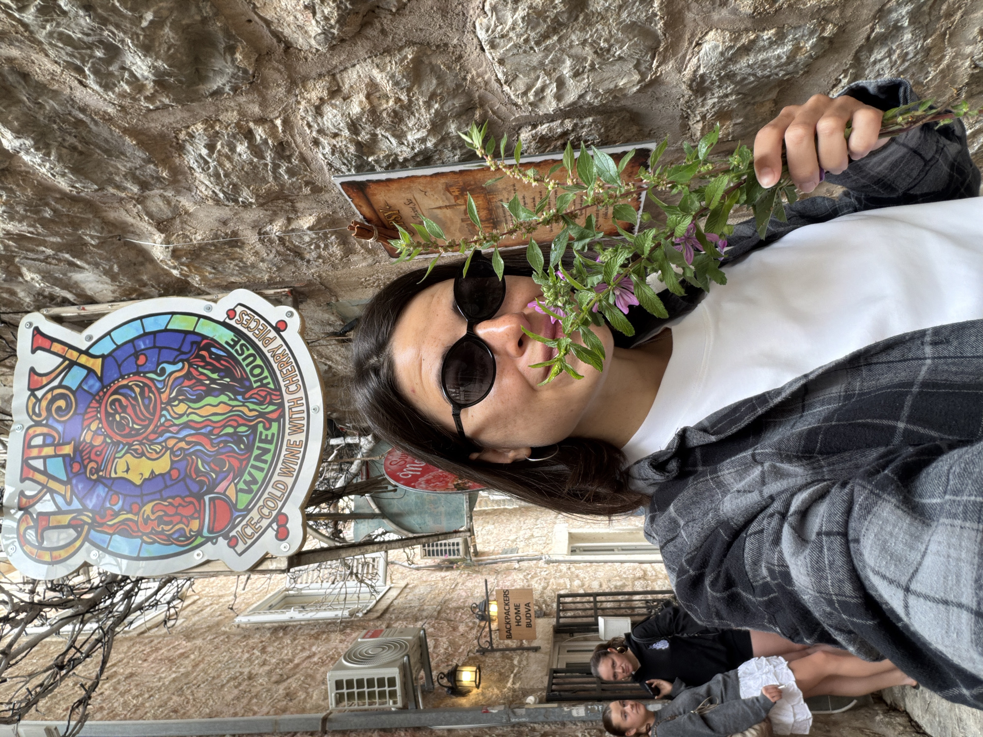 The scene is set in a narrow street of Budva, Montenegro, characterized by the old stone buildings and overhead signs. The main subject of the photo is a person wearing round sunglasses, a white t-shirt, and a gray plaid jacket, who is holding a sprig of greenery and small purple flowers near their face.
In the background, signs hang above the street. One large sign reads "TAPS HOUSE ICE-COLD WINE WITH CHERRY PIECES," featuring a colorful design of a face. Another sign says "BACKPACKERS HOME BUDVA" and is located near an exterior wall lamp. Below, a group of people are seated or lying down, one of whom is looking directly at the camera. An air conditioning unit is visible on the wall of the building. The perspective of the photo is slightly angled.