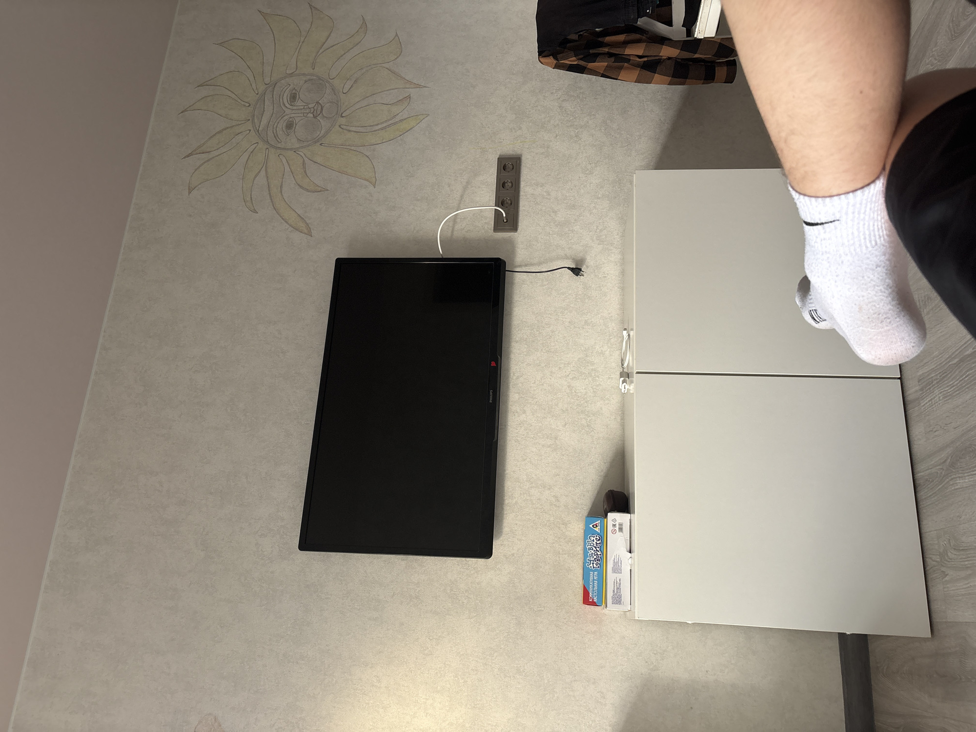 The image depicts an indoor scene, likely a bedroom or living area, with a minimalist aesthetic. A flat-screen television is mounted on a textured, light-colored wall adorned with a yellow sun motif. Below the television, a white, two-door cabinet stands on a wooden floor. On top of the cabinet, there are two boxes, one blue and white, and a small dark object. A white cable connects to a power strip with three outlets mounted on the wall. To the right of the power strip, a brown and black plaid shirt is hung on a rack. In the foreground, a person's legs, wearing white socks, are crossed, suggesting a relaxed posture. The lighting appears to be natural, coming from an unseen source.