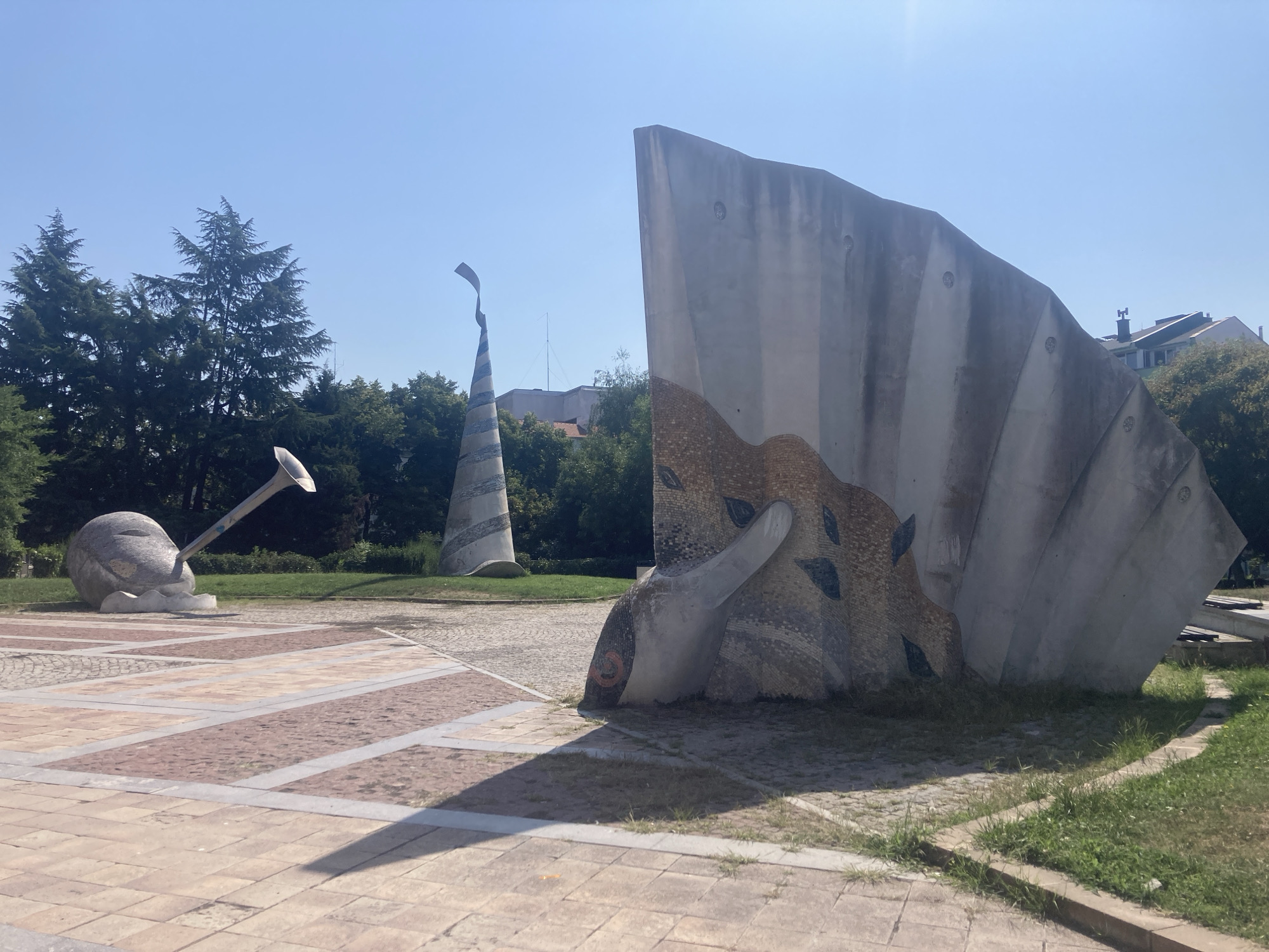 The image captures a sunny day in the Sea Garden of Burgas, Bulgaria, featuring a collection of abstract sculptures. In the foreground, a large, grey, curved structure with textured mosaic detailing dominates the right side of the frame. Its surface is imprinted with circular patterns and features an orange circular motif near its base. To the left, a rounded, stone-like sculpture with a metallic horn protruding from it rests on a base, and further in the background, a tall, conical sculpture with a striped pattern and a fluttering ribbon at its apex stands against a clear blue sky. The sculptures are situated on paved ground and surrounded by lush green grass and trees. The overall scene depicts an outdoor public art installation on a bright day.