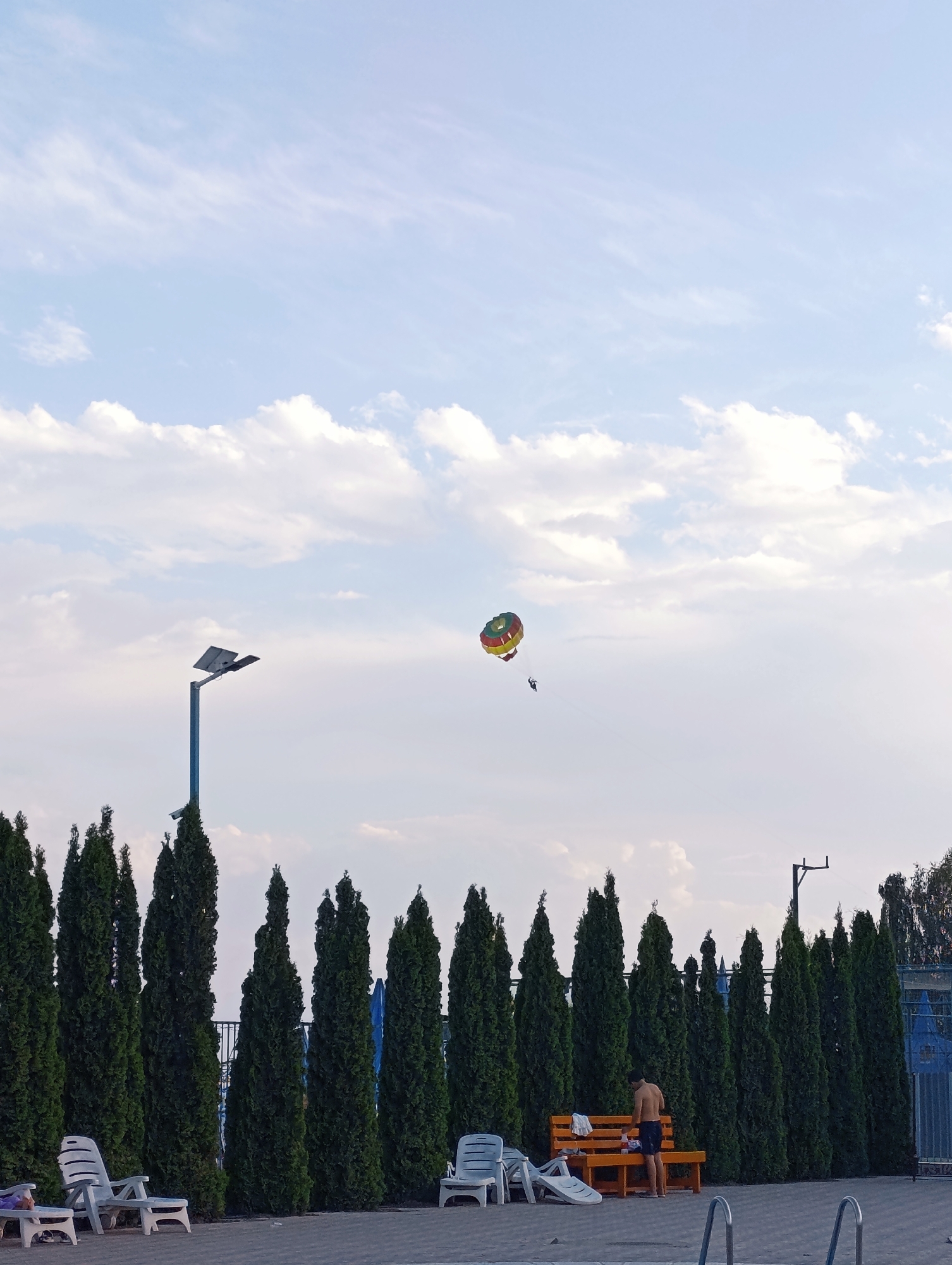 The image shows a clear sky with scattered clouds above a row of tall, dark green evergreen trees. A parasailer is visible in the sky, suspended beneath a colorful parachute. In the foreground, a man wearing dark shorts is standing next to a wooden bench. There are several empty lounge chairs and a swimming pool area. The scene appears to be an outdoor recreational area, potentially a resort or park, with elements suggesting a warm climate.