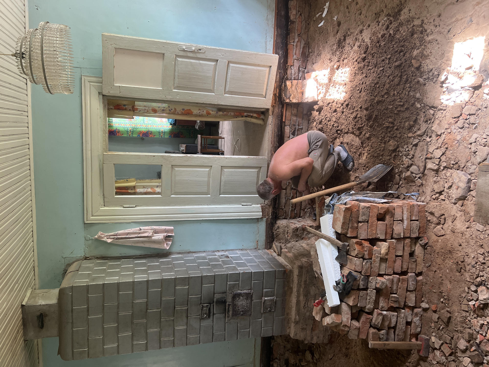 A man is kneeling on the floor of a room undergoing renovation in Бучацінскі сельскі Савет, Belarus. He is shirtless and wearing grey sweatpants and blue sandals. The floor is covered in dirt, debris, and broken bricks. A pile of red bricks is stacked to his left, with a shovel and white foam sheets on top. To his right, another shovel leans against the wall.
The room has pale blue walls and a white paneled ceiling. A large, grey tiled stove with decorative metal inserts stands prominently on the left side of the image, built on a brick base. A pink patterned garment hangs on a hook to the right of the stove. In the background, a set of double doors leads to another room, visible through the open doors are colorful curtains and a chair. A multi-tiered crystal chandelier hangs from the ceiling above the doors. The room appears to be part of a residential building.