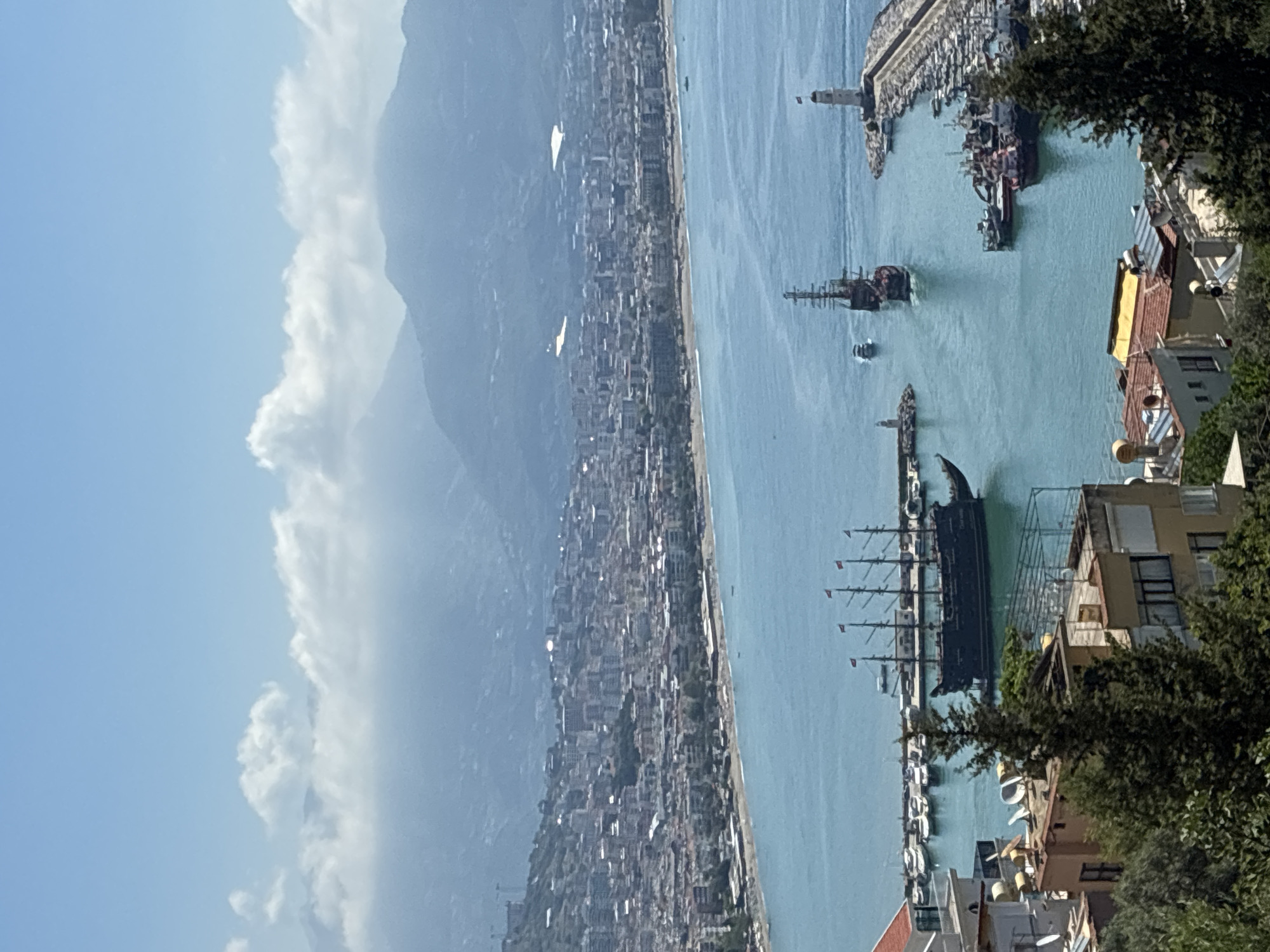 This media file captures a panoramic, high-angle view of the coastal city of Alanya, Turkey, overlooking its harbor and a long stretch of coastline, with mountains in the background under a partly cloudy sky. The image appears to be taken during daylight hours with bright, clear conditions.
In the foreground, the edge of a cliff or hill is visible on the right, covered in dense green foliage from trees. Peeking through the trees are rooftops and parts of buildings, some with satellite dishes, suggesting a residential or commercial area.
The midground is dominated by the calm, turquoise waters of a harbor. Several boats are present, most notably two large, multi-masted vessels designed to resemble old pirate ships, one of which appears to be moving or just arriving/departing. Numerous smaller boats and yachts are docked along the piers. A distinct white lighthouse stands at the entrance of the harbor on a breakwater. Beyond the harbor, a long, sandy beach or coastline extends along the sea.
Further in the midground and extending into the background is the dense urban sprawl of Alanya, with countless buildings stretching along the coast. The architecture appears typical of a modern coastal town, blending into the landscape.
In the background, majestic mountains rise, their peaks partially obscured by a hazy atmosphere and scattered white clouds. The sky above is a clear, light blue, with more defined white clouds drifting across it. No discernible text is visible within the image at this resolution. The overall scene depicts a vibrant coastal city with a busy harbor, nestled between the sea and mountains.