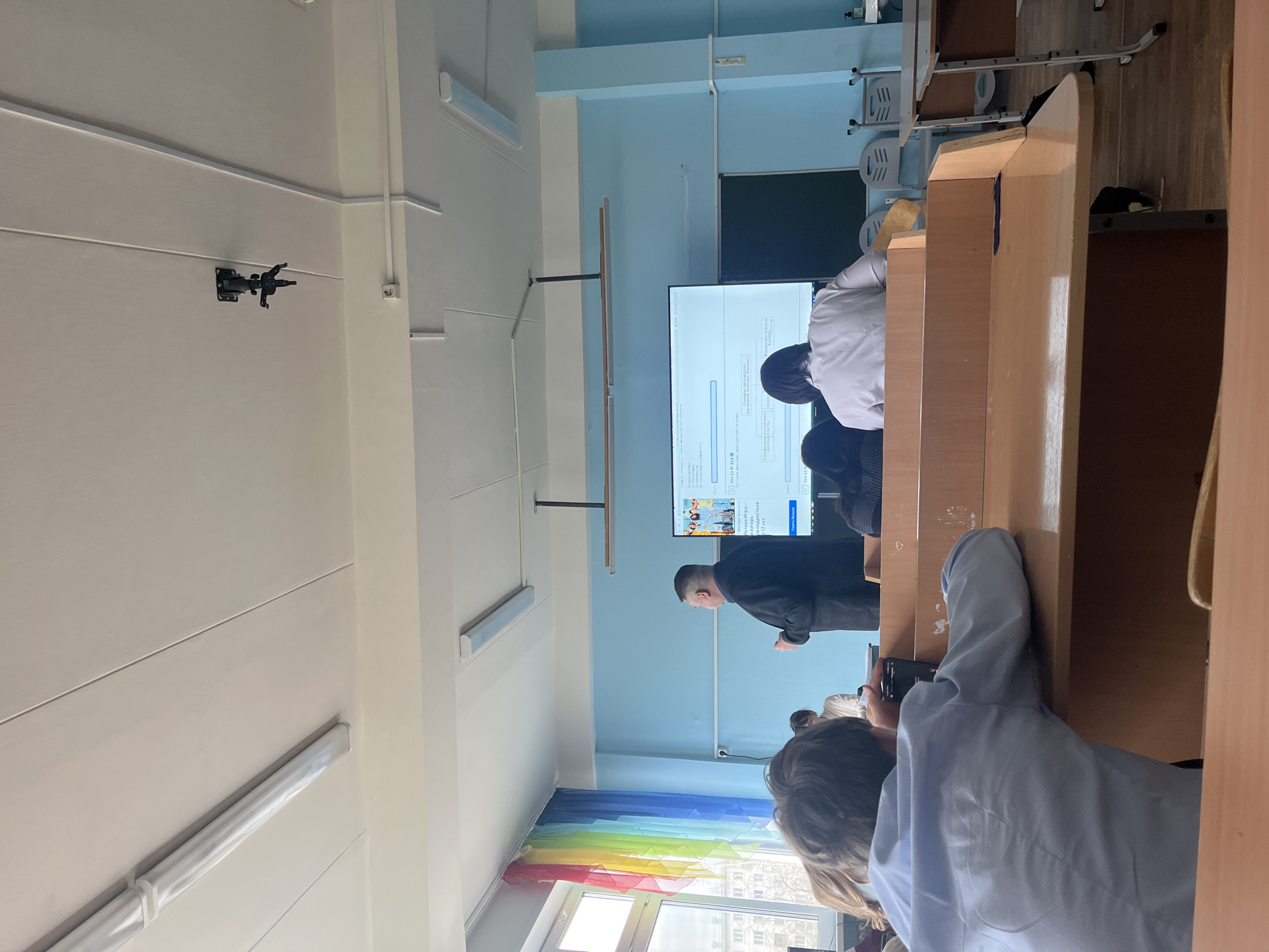 The image shows a classroom in what appears to be Chaikovsky, Russia, based on the Cyrillic text on the display. A teacher is standing near a large display screen, presumably lecturing or presenting information to students. Students are seated at desks, with one student in the foreground possibly recording or photographing the screen. The classroom has a light blue wall color, fluorescent lighting, and a window with a rainbow decoration. Desks are arranged in rows. A camera mount is visible on the wall near the ceiling.