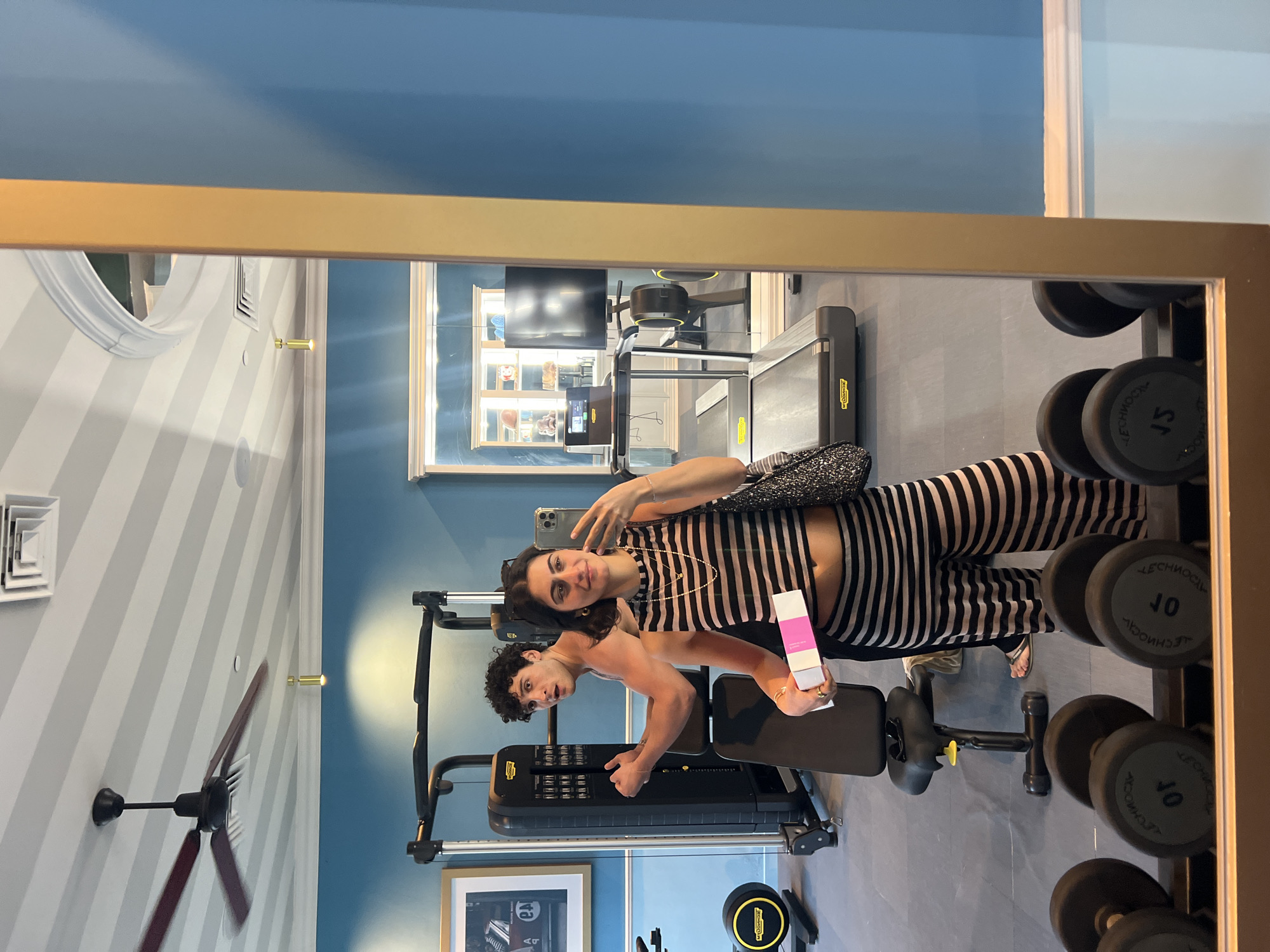 In Santa Eulària des Riu, Spain, a photo is taken in a gym or fitness center as reflected in a large, gold-framed mirror. A woman in striped workout attire holds up a mobile phone, taking a selfie. Alongside her, a shirtless man with curly hair flexes his arm muscles and gives a thumbs-up gesture. He holds a white and pink box in his other hand. The gym is well-equipped with exercise machinery, including a cable-based machine, dumbbells, and a treadmill. The walls are a mix of blue and white striped, and there is a ceiling fan and framed artwork visible.