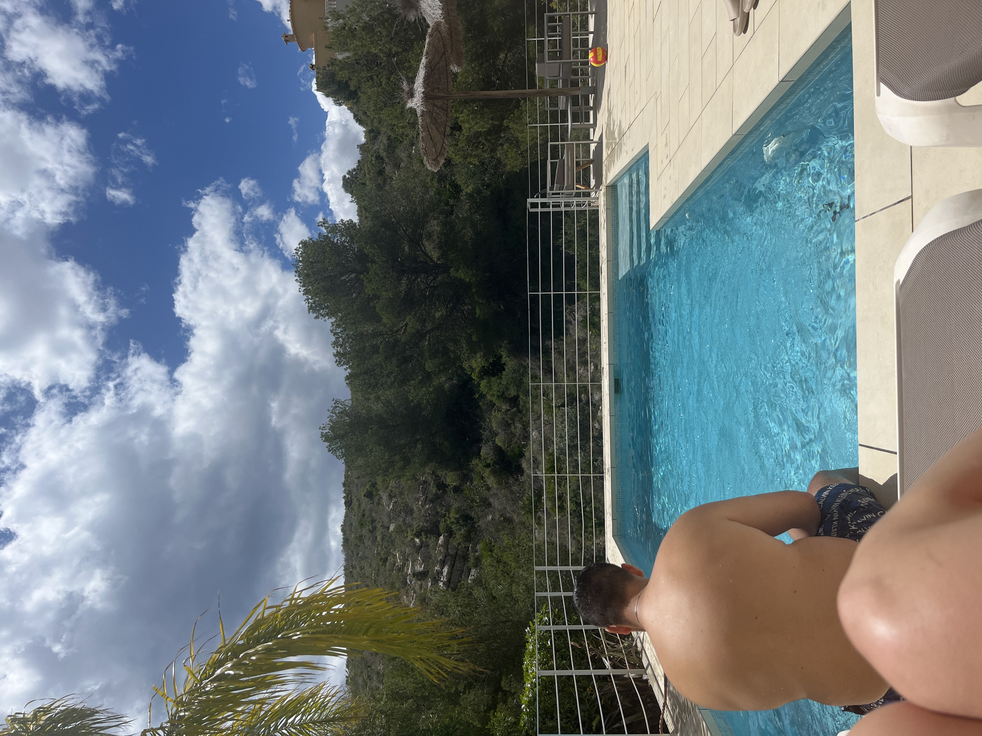 The image shows a shirtless man sitting by a blue swimming pool, with green trees and a mountain in the background, under a partially cloudy sky. The scene is likely located in El Poble Nou de Benitatxell / Benitachell, Spain. The pool is bordered by light-colored tiles and a white fence. A palm tree is visible in the lower left corner. There are sun loungers near the pool.