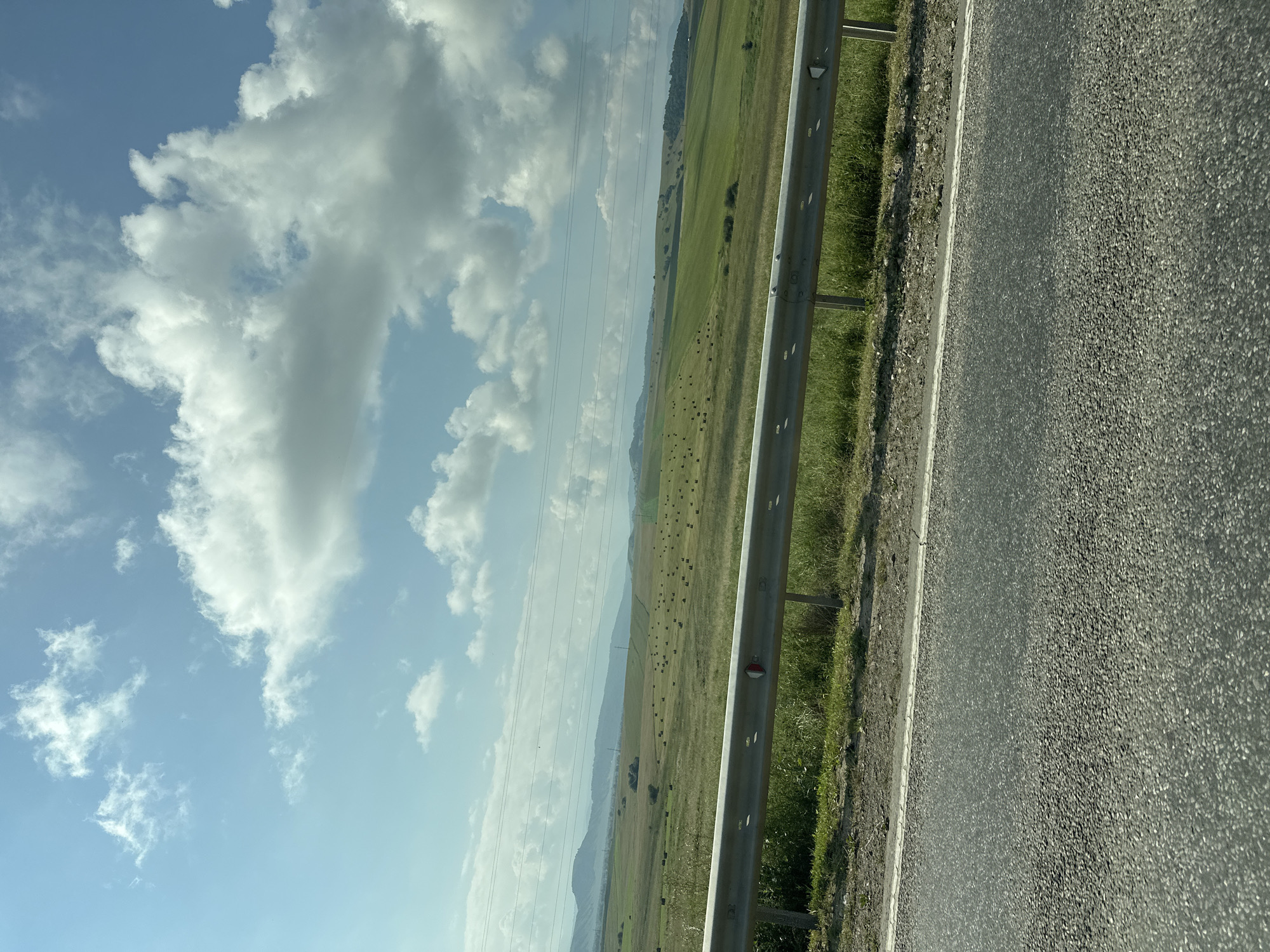 The image captures a rural landscape under a partly cloudy sky. In the foreground, a grey asphalt road stretches across the bottom of the frame, bordered by a narrow strip of dry earth and then a metal guardrail. Beyond the guardrail, a grassy field slopes gently upwards, dotted with numerous dark, circular hay bales. Further in the distance, rolling hills covered in green grass and scattered trees recede towards a hazy horizon. Power lines are visible stretching horizontally across the middle ground. The sky above is a clear blue with scattered white, puffy clouds, some of which are illuminated by sunlight. The scene is devoid of any people or animals. This is a view of a rural landscape, likely in the vicinity of Zelenchukskaya, Russia, characterized by agricultural fields and natural terrain.