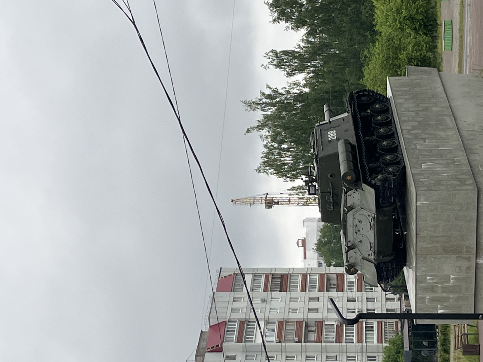 The image shows a T-34 tank mounted on a concrete plinth, typical of a World War II memorial. The tank is green and has the number "288" painted on the turret. A red star is visible on the side of the tank. The tank is viewed from a low angle. In the background is an apartment building and a construction crane. A set of power lines stretches across the sky. The sky is overcast.
Based on the visible buildings and the presence of the T-34 tank memorial, this photo was likely taken in the city of Chaikovsky, Perm Krai, Russia. The image has been rotated 90 degrees clockwise.