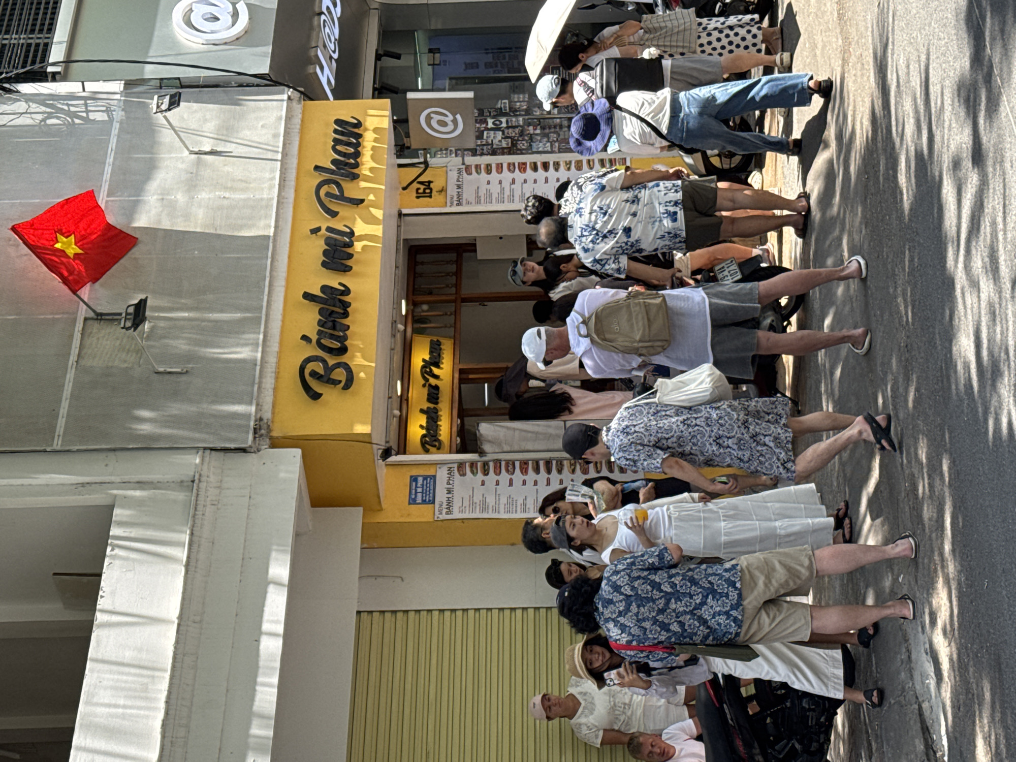 This image depicts a street scene in Nha Trang, Vietnam, likely outside a food establishment. A bright yellow facade with "Bánh mì Phan" written in cursive black lettering dominates the center of the frame. Above this sign, a Vietnamese flag is displayed on a pole attached to the building. To the right of the entrance, a sign with the number "164" and an "@" symbol is visible. A menu board for "BÁNH MÌ PHAN" is also mounted on the wall near the entrance.
A group of people, appearing to be customers, are gathered on the sidewalk in front of the establishment. They are of various ages and genders, dressed in casual summer clothing. Some are looking at the menu, while others appear to be waiting or talking. One woman holds a drink, and a man in the foreground wears a backpack. The street in front of the building is paved and shows shadows cast by trees, suggesting a sunny day. A motorcycle is parked to the left, and to the far right, a person is holding a white umbrella. The building itself has a textured, light-colored exterior above the yellow sign. The overall atmosphere suggests a busy urban environment with commercial activity.
