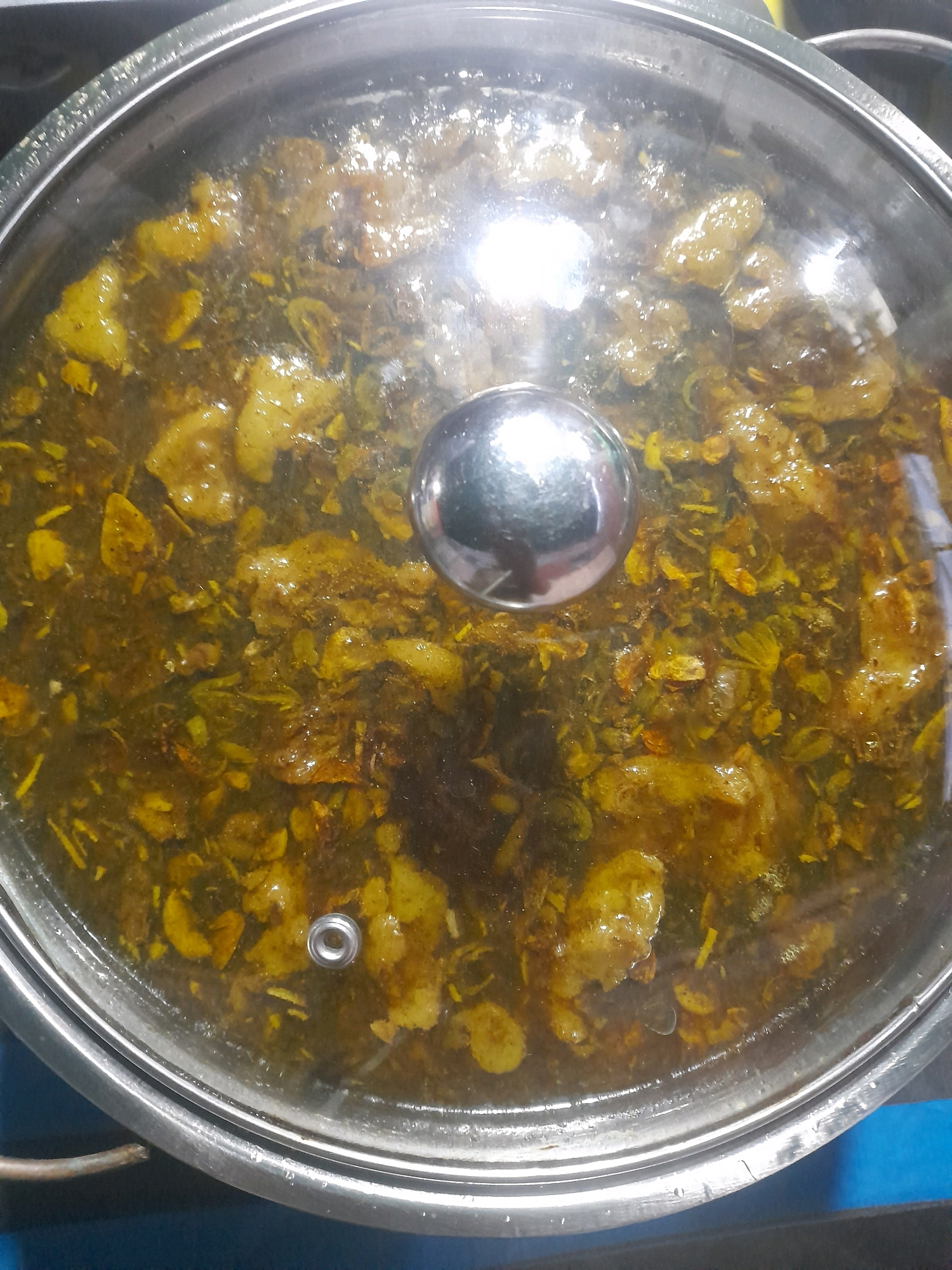 In a kitchen in Mlaras, Indonesia, a pot with a clear glass lid sits on a stove. The pot contains a simmering curry with pieces of meat and vegetables in a rich, yellowish-brown sauce. The lid reflects the overhead lights, obscuring some of the details of the food. A stainless steel handle is visible on the lid. The pot is silver and has a rounded rim. The background is blurred, but suggests a kitchen environment with blue and black surfaces.