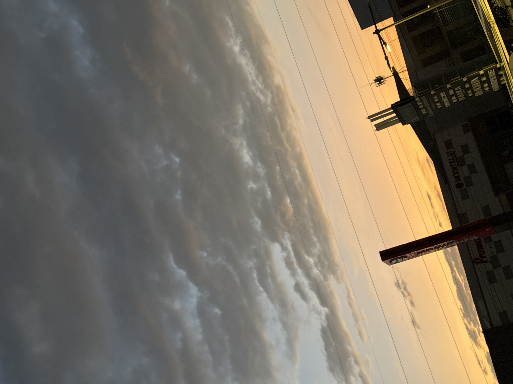 The image displays an urban scene at either sunrise or sunset in Puebla, Mexico, rotated ninety degrees counter-clockwise. In its correct orientation, the sky occupies the upper portion, showcasing a dramatic contrast between dark, heavy clouds on the left and a radiant, golden-orange hue on the right. Several power lines cut across the sky, stretching diagonally from the lower left to the upper right.
In the lower right section, commercial buildings are visible, appearing as dark silhouettes against the bright horizon. A street light is illuminated, suggesting dusk or dawn. Several business signs are discernible on the buildings. One sign clearly reads "MODATELAS" within a checkered design. Another partially visible sign shows text that appears to include "Snobove," "BAIKA," and "WAGHA." A prominent red rectangular sign is also present, with the word "Proma" partially visible.
No people or specific activities are depicted, allowing the focus to remain on the interplay between the urban landscape and the impressive sky. The heavy clouds suggest dynamic weather or a visually intense atmospheric condition. The overall mood is striking, capturing a moment of transition in an urban setting.