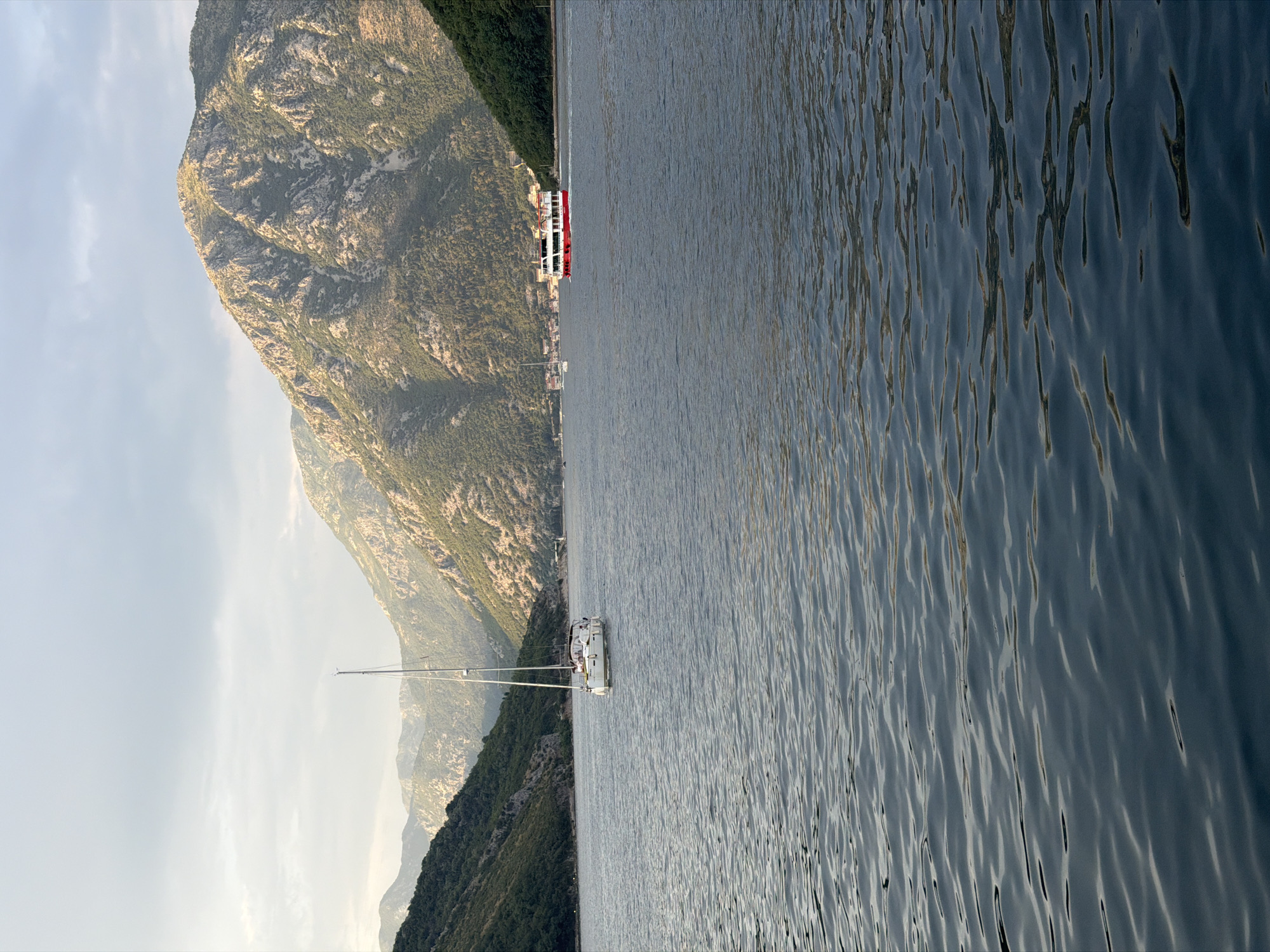This is a landscape photograph of a bay with a sailboat and a ferry. The foreground is dominated by the dark blue, rippling water of the bay. In the middle ground, a white sailboat is visible on the left, with several people on deck. Further to the right, a red and white ferry is traveling across the water. The background features steep, tree-covered mountains that flank the bay. The sky is a hazy, pale blue with some clouds, and the sunlight appears to be soft, suggesting it might be early morning or late afternoon. The location is Lepetane, Montenegro, as indicated by the distinctive mountainous terrain and the bay setting, characteristic of the Bay of Kotor.