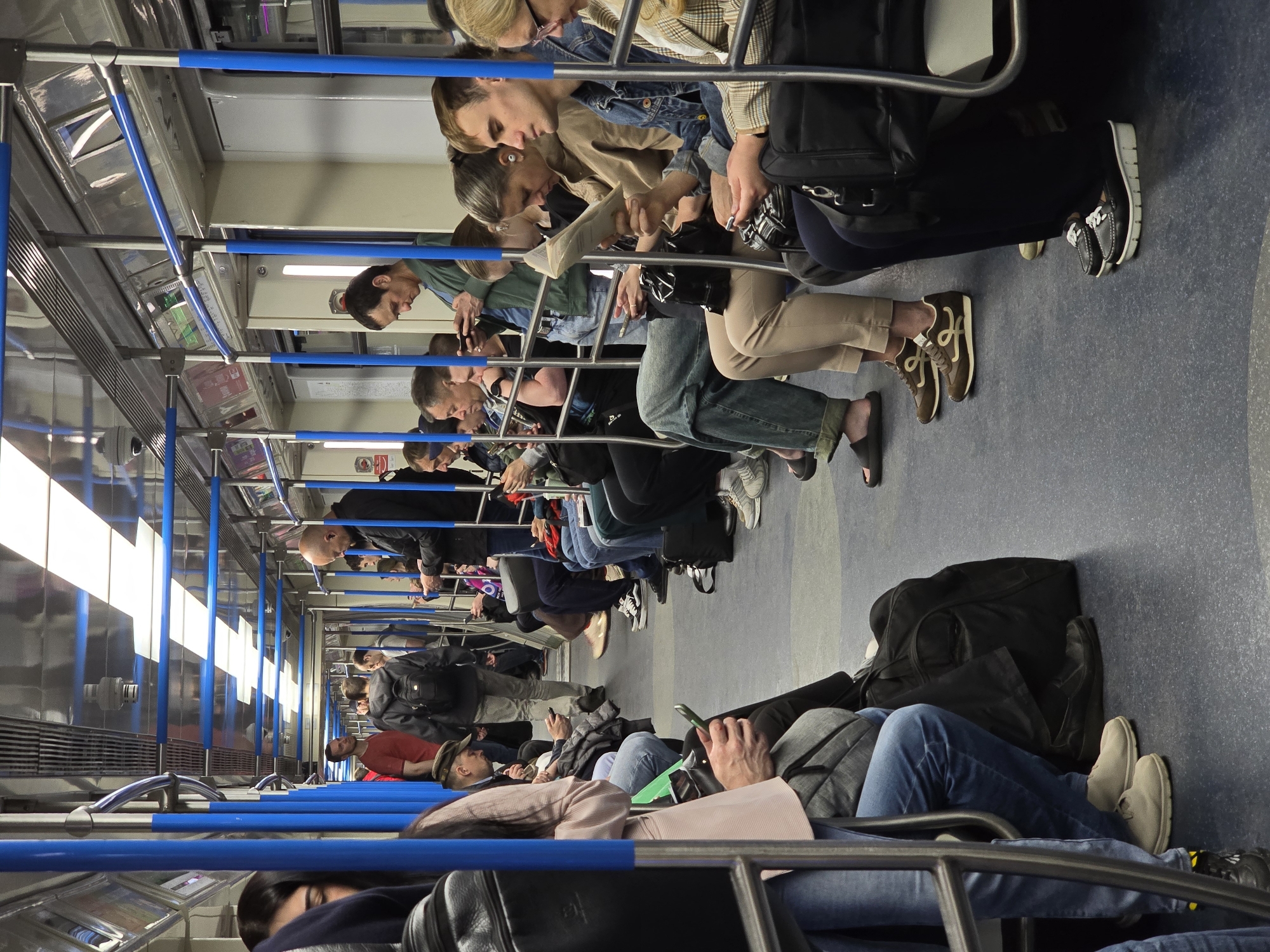 The image depicts the interior of a subway car in an unknown city. The scene is populated with seated passengers, most of whom are looking down at their phones. The car features rows of seating along the sides, with blue handrails running along the ceiling for standing passengers. The subway car appears clean and well-lit, with white ceiling panels and stainless steel or metallic walls. Passengers are dressed in casual clothing. Luggage, including backpacks and bags, is visible on the floor and on laps. The overall atmosphere is one of quiet, focused activity as people commute, likely to or from work or other daily activities.