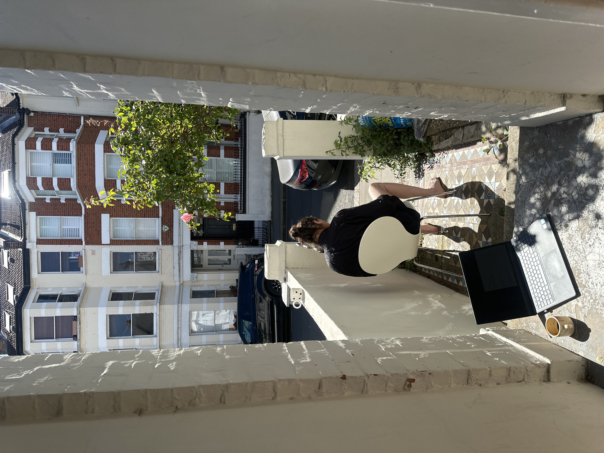 This image captures an outdoor scene from a slightly elevated perspective, possibly a front garden or patio, overlooking a street in a residential area of London, United Kingdom.
In the foreground, a person is seated on a contemporary white chair with their back to the viewer. They are wearing a dark top and shorts, with their hair tied up. To their right, resting on a low white wall, is an open laptop with a dark screen and a light-colored mug. The ground beneath the chair features decorative, patterned tiled paving, which transitions to a more worn concrete surface further into the scene. Both surfaces are dappled with strong sunlight and shadows, indicating a bright, sunny day, likely in the morning or late afternoon.
Beyond the immediate foreground, a narrow street is visible, with a dark car parked on it. Across the street, a row of traditional brick terraced houses lines the opposite side. These multi-story homes feature white window frames, some with bay windows and white architectural details that stand out against the red brick. A green leafy tree partially obscures the view of the houses and street. The architectural style, street layout, and presence of terraced housing are characteristic of London's urban environment. No distinct, legible text is visible in the image.