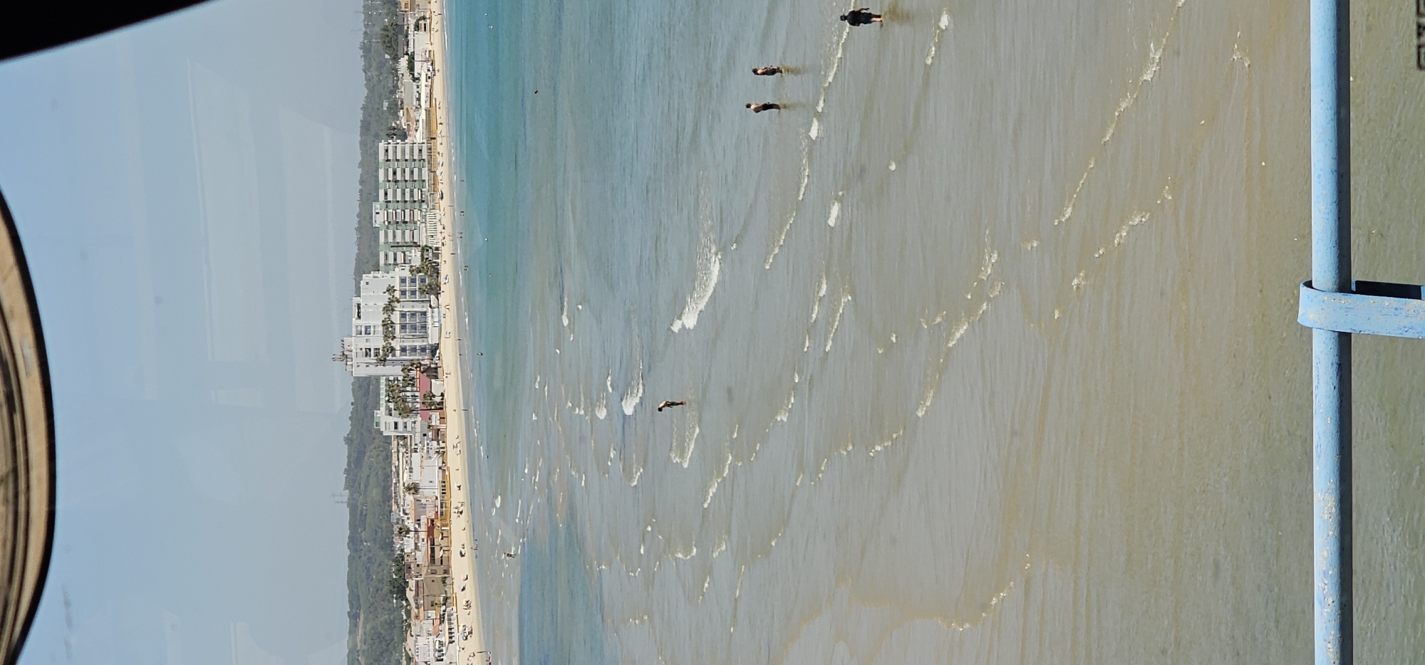 This aerial-like shot captures a vibrant beach scene from an elevated perspective, likely from a window or balcony, with a portion of a round metallic frame visible on the left and a blue railing on the right. The image is oriented vertically but depicts a horizontal landscape.
The expansive sandy beach, dotted with numerous beachgoers, stretches along the coastline, bordered by a line of multi-story buildings, including hotels and apartments, nestled amidst green foliage. The buildings vary in architectural style, with some appearing modern with multiple balconies, while others have simpler designs.
The sea dominates the central and right portions of the frame, showcasing a range of blues and greenish-brown hues. This color variation suggests clearer water closer to the shore and perhaps sandy sediment stirred up further out. Small, gentle waves break rhythmically across the water's surface. Several individuals are visible in the water; some are standing in the shallower areas, while others are further out, possibly swimming or wading. At least four distinct figures can be seen dispersed in the water.
The scene conveys a bright, clear daytime setting, indicative of good weather for beach activities. There are no clouds visible in the sky, which appears a light, hazy blue. No specific text or signs are discernible in the image. The overall impression is one of leisure and activity on a popular coastal destination, possibly in Algeciras, Spain, given the provided context.