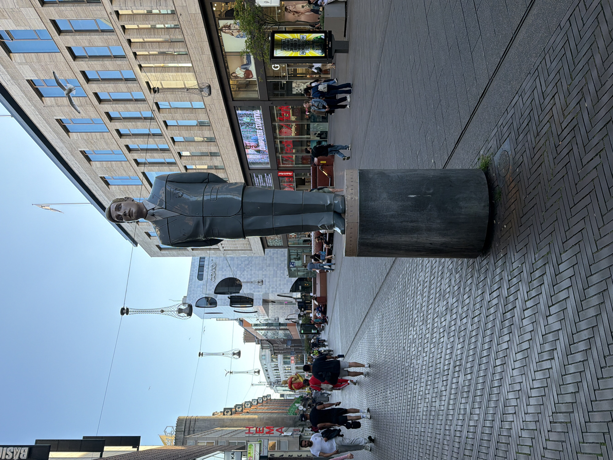 A statue of a man in a suit stands on a pedestal on a city street. The statue is positioned in the foreground, facing forward. Behind the statue, the street is paved with cobblestones on the left and a darker, smoother pavement on the right. Buildings line both sides of the street. On the right, a large modern building with many windows is visible, with advertisements displayed in its lower level. On the left, more traditional-looking buildings with shops are present. People are walking on the street, some in groups and some individually. Some are engaged in shopping, while others are simply passing through. There are also bicycles visible, suggesting that cycling is a common mode of transport in this area. Overhead, decorative light fixtures hang from wires. The sky is clear and blue, indicating a pleasant day. This scene is located on the Grote Marktstraat in The Hague, Netherlands, a well-known shopping street in the city center.
