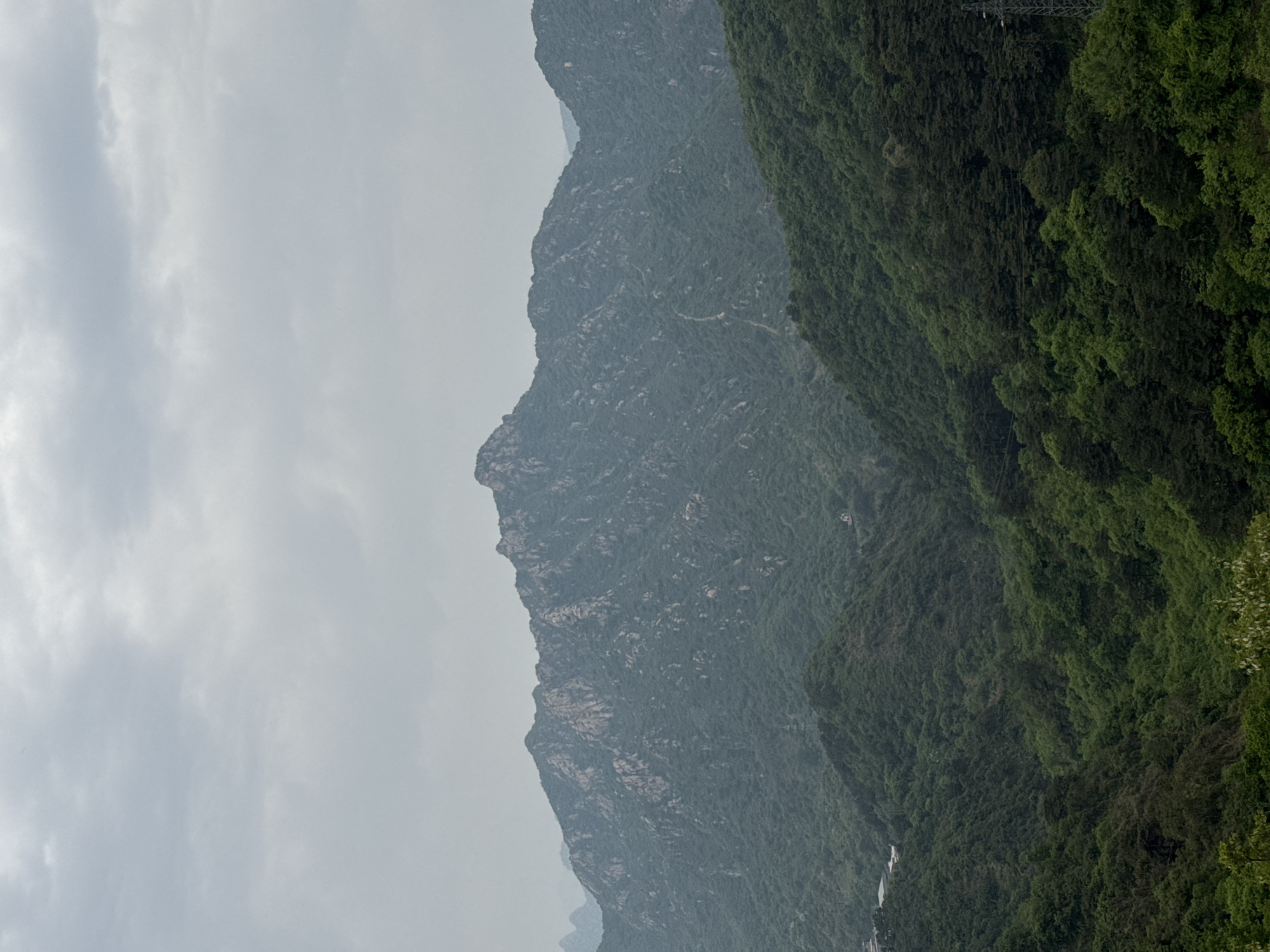 The image captures a mountainous landscape in the Huairou District of China, under an overcast sky. The composition is dominated by two primary mountain ridges. The mountain on the right is densely vegetated with lush, green foliage, extending from the foreground towards the top of the frame. The second mountain, positioned to the left, presents a rugged, rocky terrain with sparse vegetation, exhibiting a greyish hue due to the weather conditions. The sky, visible on the left side, is covered with clouds, creating a muted, diffused light across the scene. At the very bottom of the image, hints of infrastructure, such as buildings or roads, can be discerned amid the dense vegetation. Power lines are visible on the right side of the image.