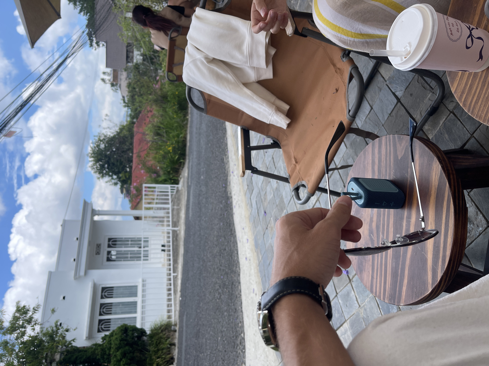 A first-person perspective photograph shows a person holding sunglasses over a small, round, wooden table in an outdoor setting. A small, dark blue speaker sits on the table. A paper cup with a straw is visible in the upper right of the frame. In the background, two people are seated in chairs next to a brown jacket. A white building with a white fence is visible across the street. The location appears to be a cafe in Da Lat, Vietnam, as indicated by the writing on the paper cup. The ground is paved with gray stones. The sky is partially cloudy.