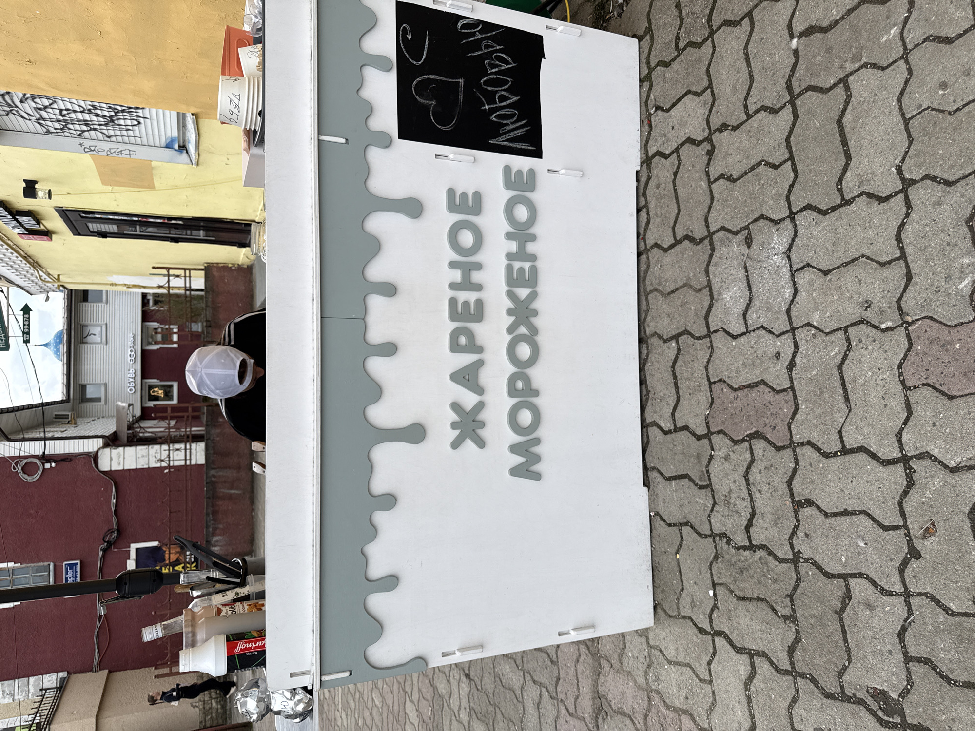 An outdoor scene in Gatchina, Russia, features a white kiosk with grey dripping accents. The kiosk has raised grey lettering that reads "ЖАРЕНОЕ МОРОЖЕНОЕ" (Fried Ice Cream). To the right, a small blackboard with chalk drawings of a heart and the word "ЛЮБОВЬЮ" (with love) is attached to the kiosk. A person wearing a white baseball cap and a dark shirt is partially visible behind the counter of the kiosk. Various bottles and containers are on the counter. The kiosk is situated on a paved walkway made of interlocking grey bricks. In the background, buildings with red and yellow facades are visible, along with street signs and other urban elements.